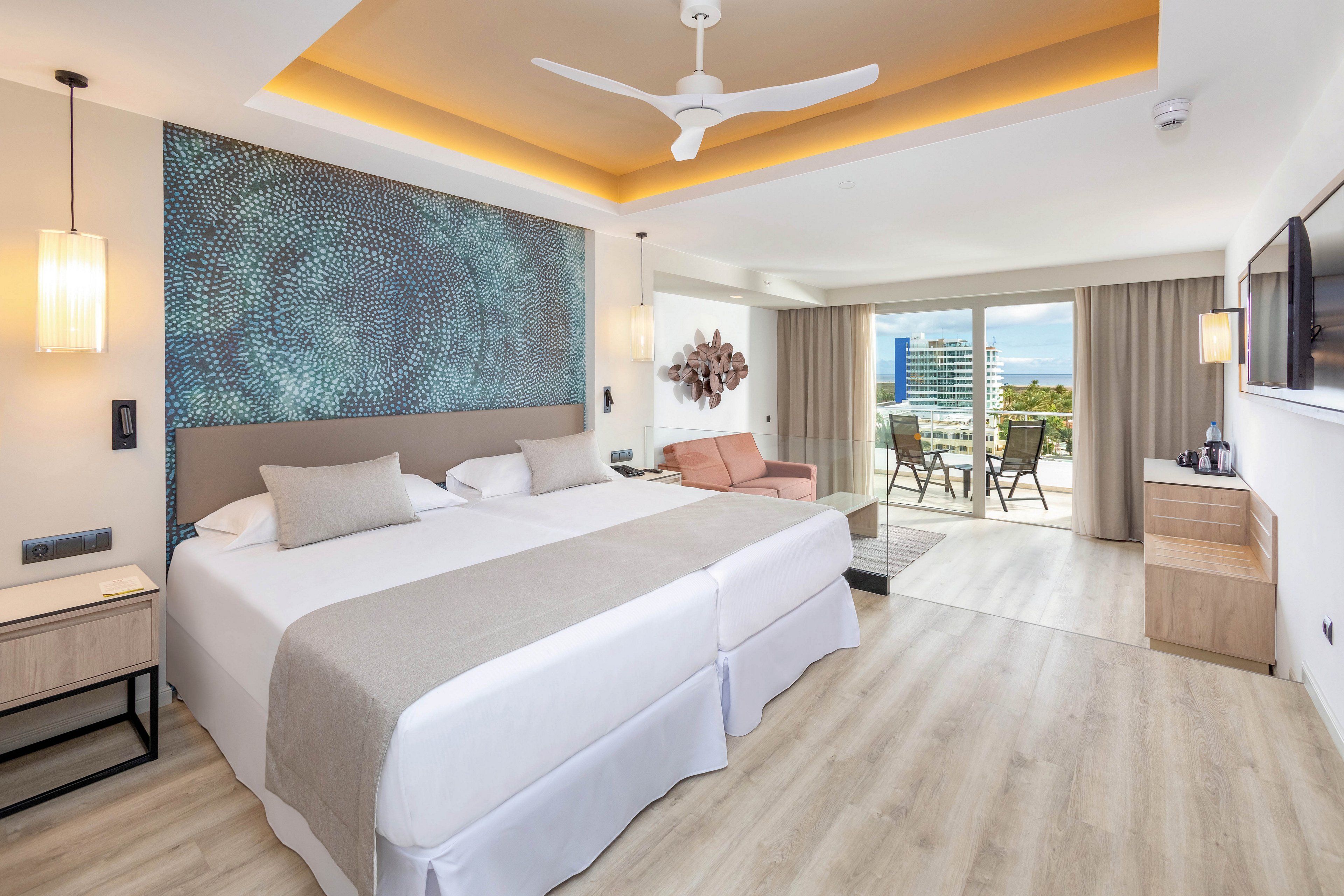 A spacious hotel room features two large beds with neutral bedding, a contemporary wall art accent, and a cozy seating area. The room is well-lit with natural light from a glass balcony door, offering a view of city buildings and greenery. Modern furnishings, a ceiling fan, and a flat-screen TV enhance the comfortable, stylish atmosphere. The palette is soft and inviting, with beige, white, and muted blue tones.