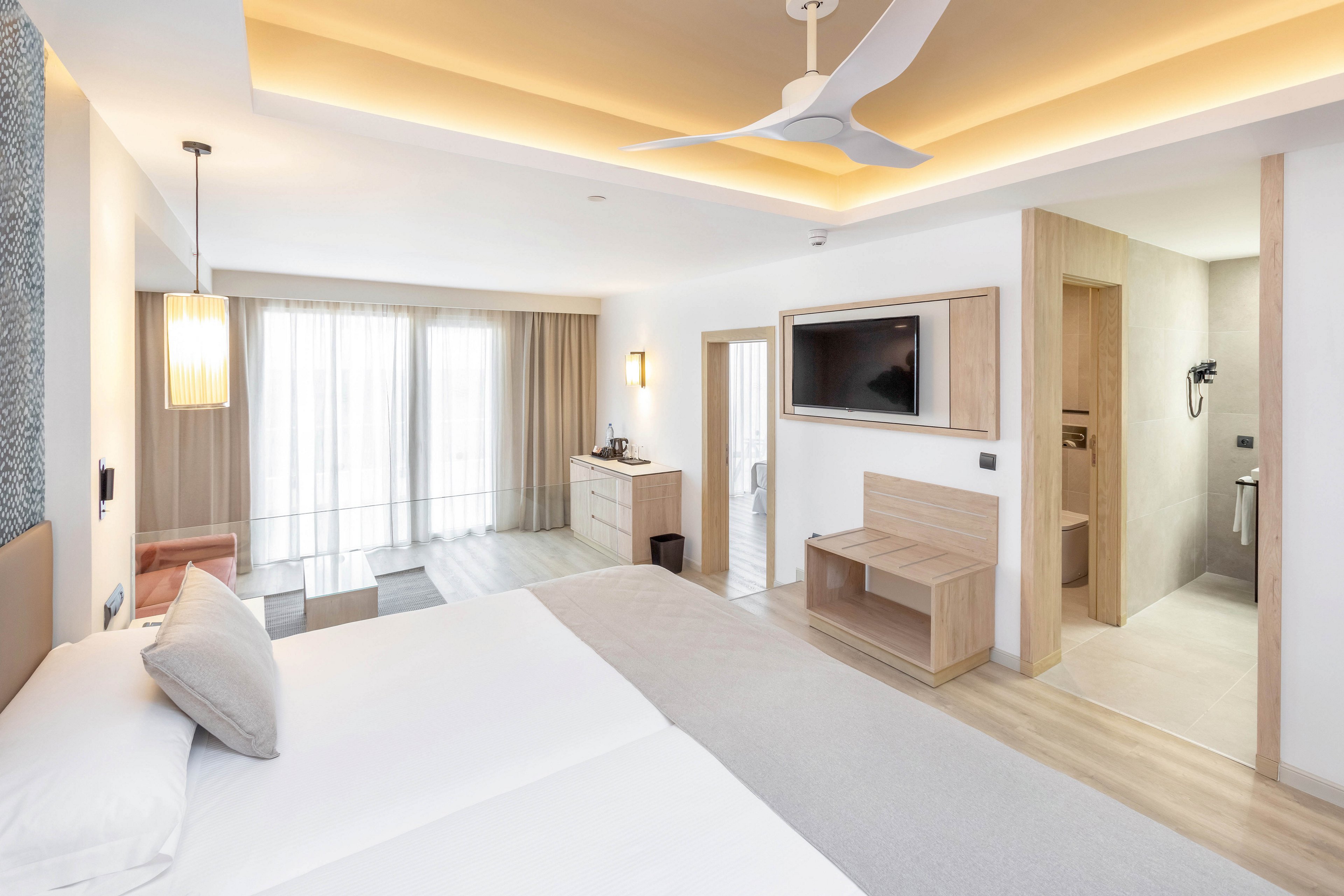 A spacious, contemporary hotel room features a large bed with crisp linens, a wall-mounted television, and a minimalist desk. The setting is bright with natural light streaming through sheer curtains, and the bathroom area is visible with sleek fixtures. The mood is calm and inviting, highlighted by soft beige and white color palette and ambient ceiling lighting.