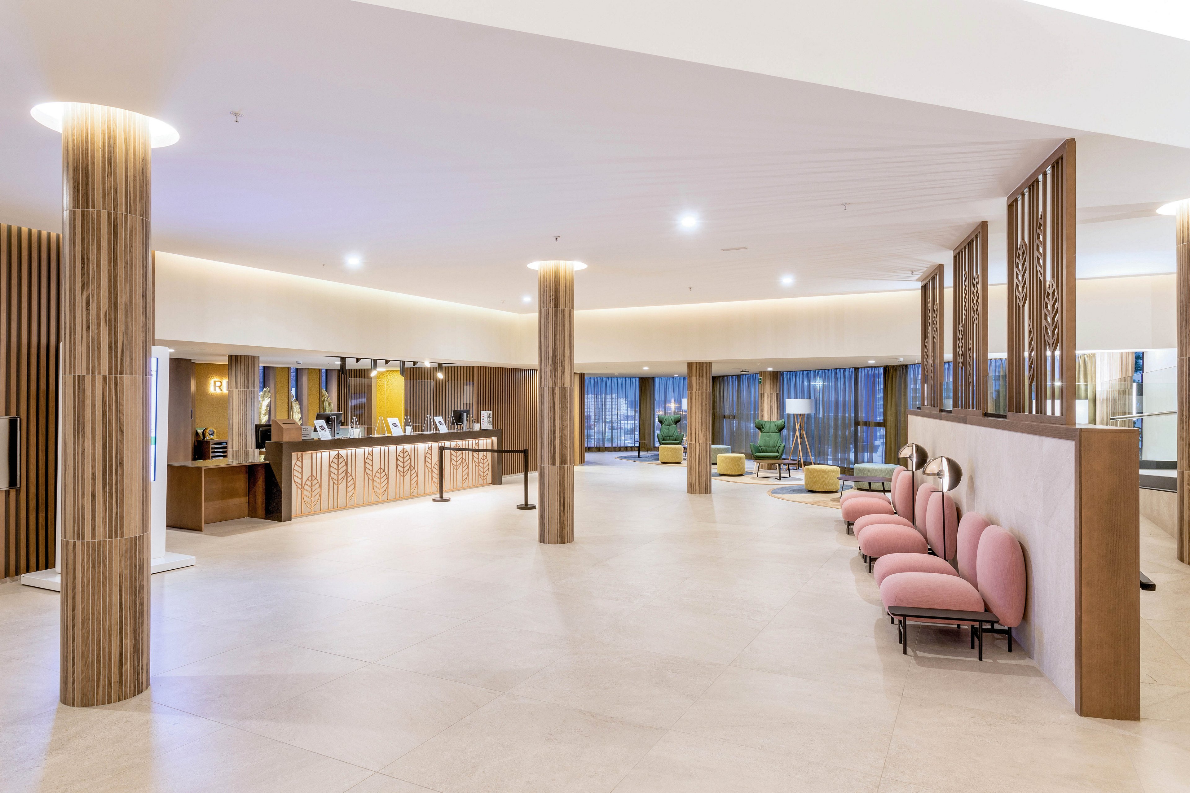 Spacious hotel lobby featuring a contemporary reception desk and a row of pink cushioned chairs. The setting is bright and airy, with wood accents and large windows providing natural light. The mood is welcoming and calm, with neutral tones and minimalist decor. No visible people or text are present in the image.