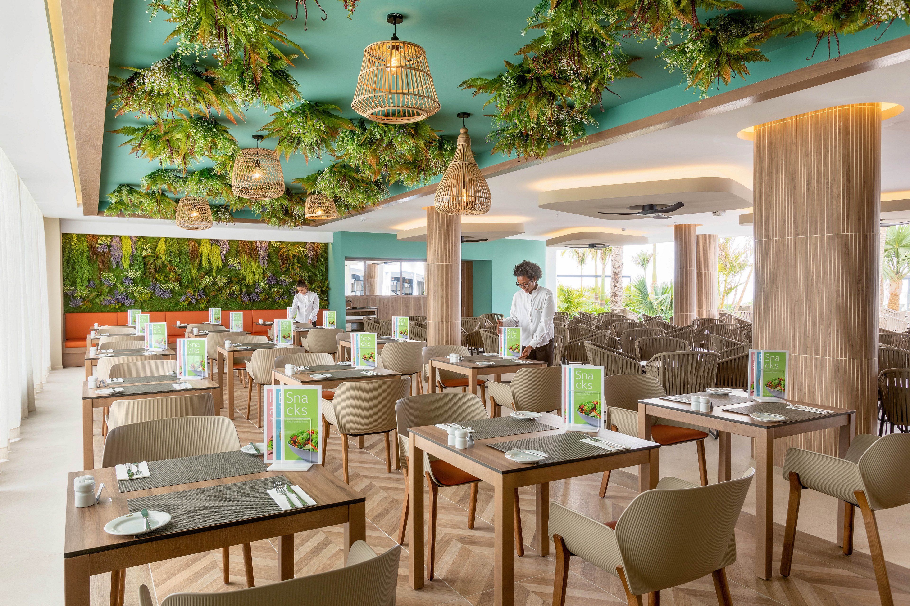 A contemporary restaurant dining area features wooden tables and beige chairs, with lush green plants adorning the ceiling and walls. Two staff members, dressed in white shirts, are preparing the tables, which display menus with visible food images and text. The space is well-lit with natural light and decorative pendant lights, creating a fresh and inviting atmosphere. No explicit numeric values or unmistakable brand names are visible.