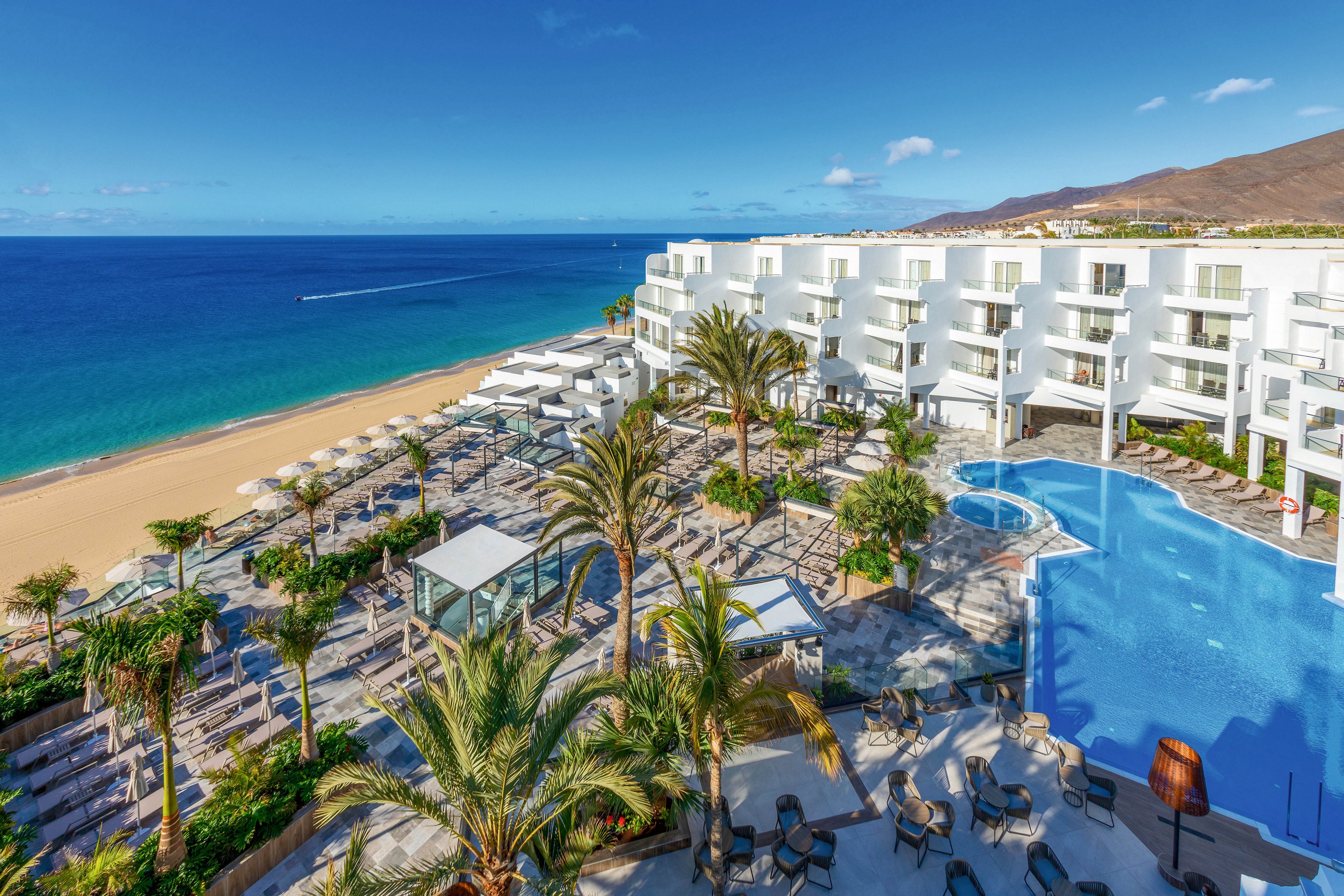 A contemporary white hotel overlooks a pristine sandy beach and turquoise ocean. The setting features a large outdoor swimming pool surrounded by palm trees, sun loungers, and umbrellas, creating a relaxing vacation atmosphere. The scene is bright and sunny, with clear skies and a tranquil mood, ideal for leisure and travel. No visible text or numbers are present in the image.