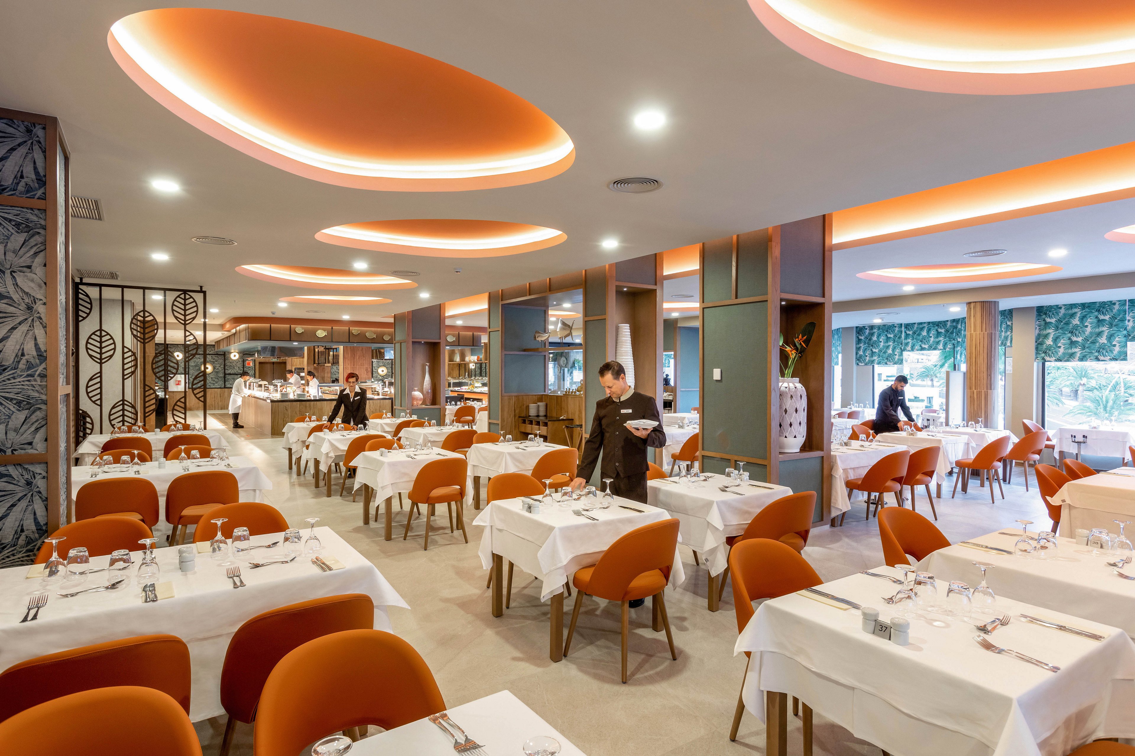 A spacious restaurant dining area features neatly arranged tables with white tablecloths and orange chairs. The ceiling has unique circular lighting fixtures emitting a warm glow, creating a contemporary and inviting atmosphere. A few staff members are seen preparing tables, and large windows provide natural light. The setting is clean and stylish, ideal for casual or formal dining.