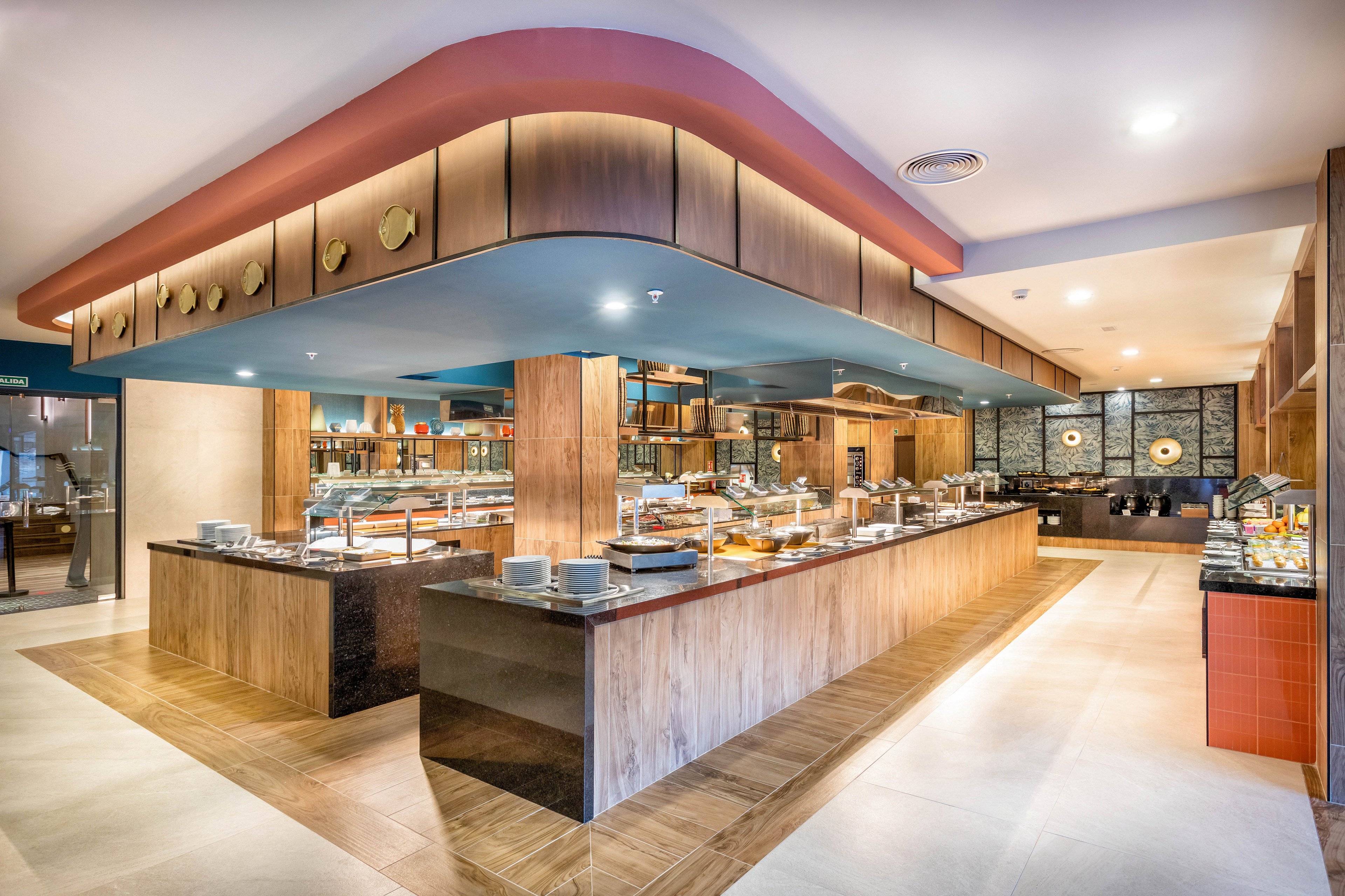 A spacious, contemporary buffet restaurant features a central island with a variety of food stations and serving dishes. The setting is bright and inviting, with wood accents, sleek countertops, and warm lighting. Plates and utensils are neatly arranged, and the overall mood is clean and welcoming. No visible text or numbers are present in the image.