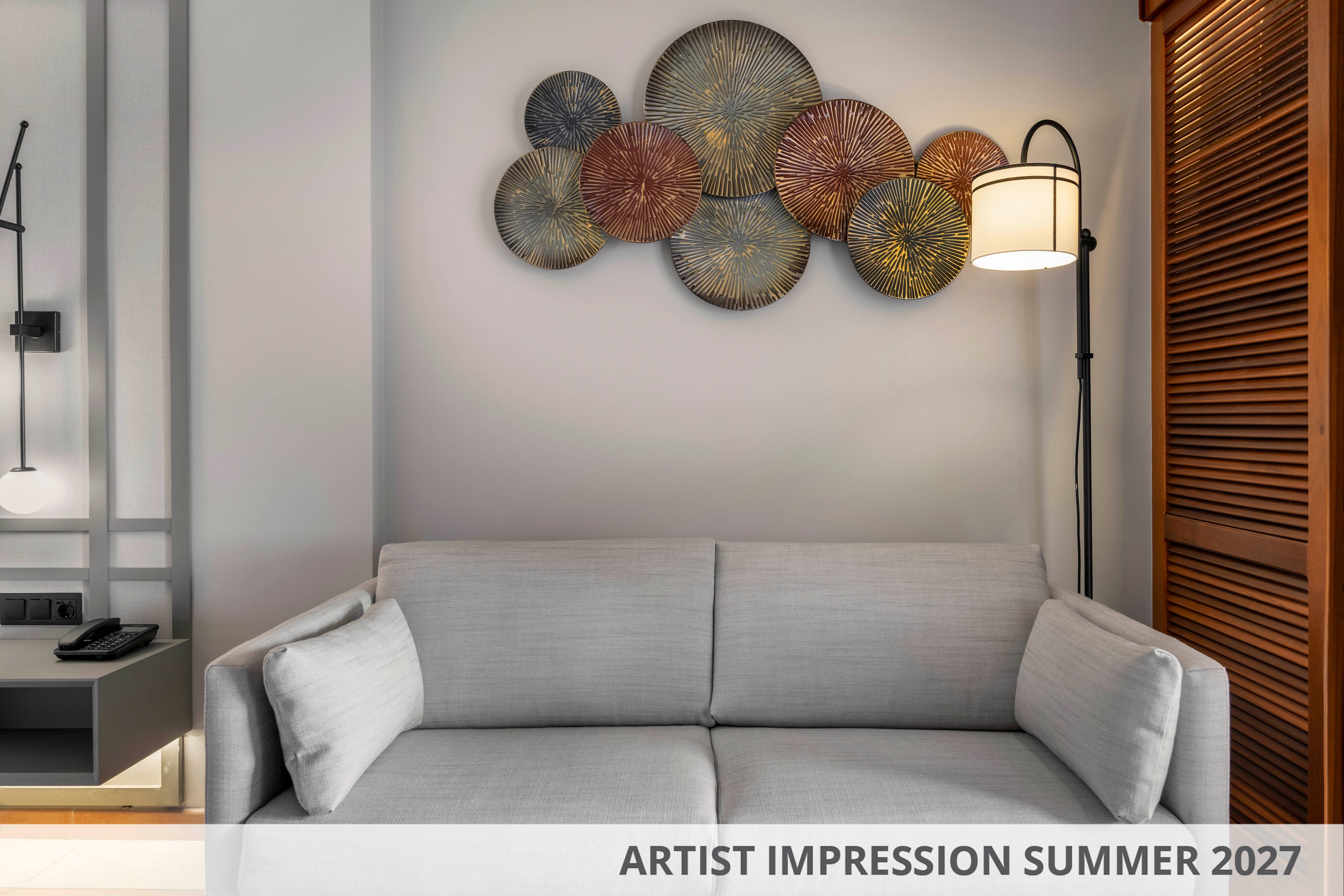 Contemporary living room interior featuring a light gray sofa against a neutral wall. Decorative circular metal wall art adds texture and visual interest above the seating area. A floor lamp stands to the right, while a built-in desk with phone and remote is visible to the left. Text at the bottom reads ARTIST IMPRESSION SUMMER 2027, indicating a conceptual design for a future space.