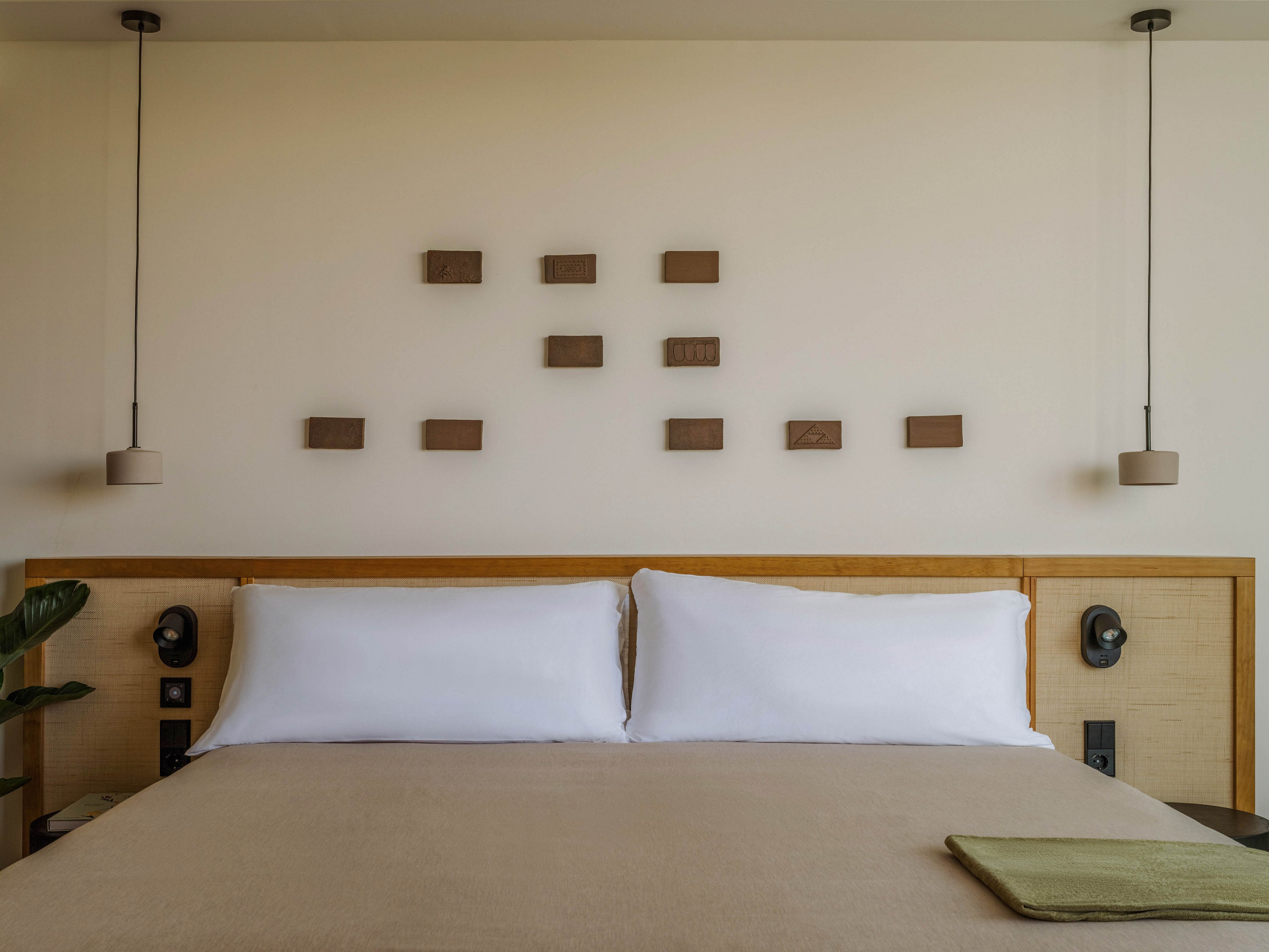 A neatly made bed with white pillows and a beige bedspread is centered against a light-colored wall in a minimalist bedroom. Small rectangular wooden art pieces are arranged in a grid pattern above the headboard. Two pendant lights hang symmetrically on either side of the bed, adding a modern touch. A folded green cloth lies on the bed, and a plant is partially visible to the side, enhancing the calm, contemporary atmosphere.