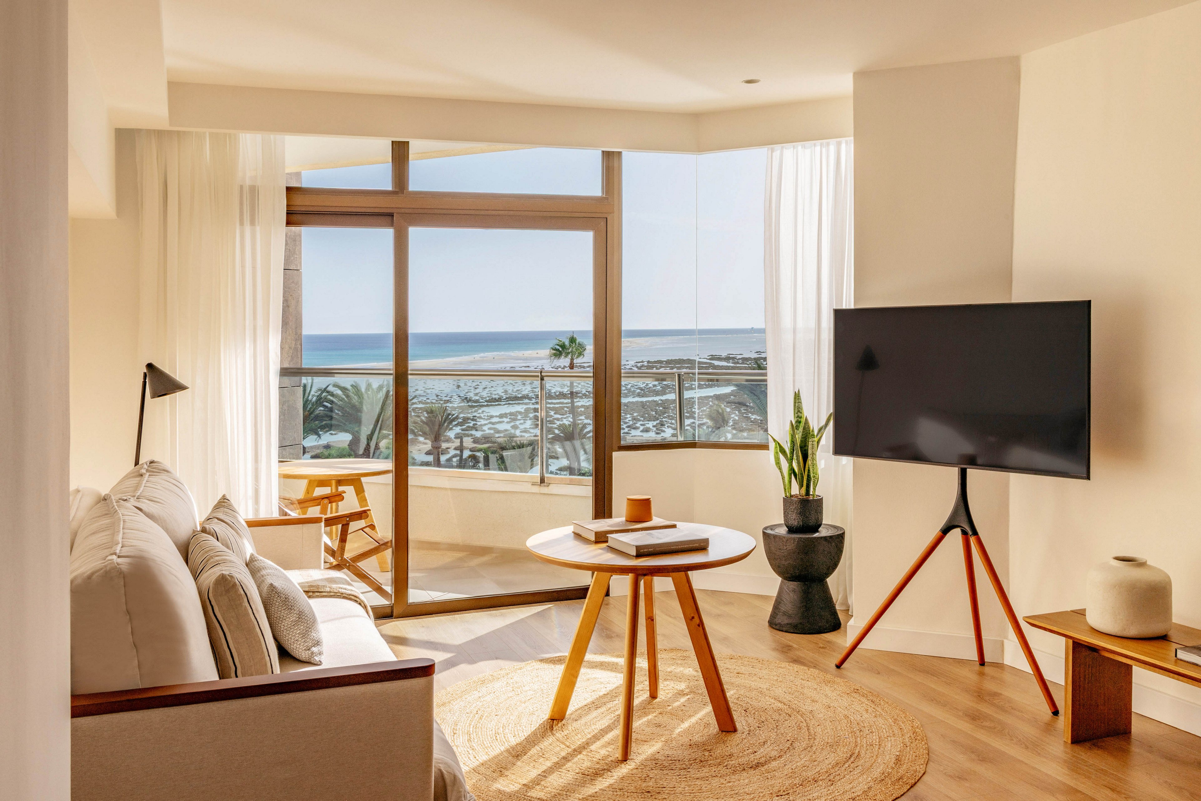 A bright modern living room features a neutral sofa and a round wooden coffee table on a woven rug. Floor-to-ceiling windows open onto a balcony overlooking a sunny beach and ocean. A flat-screen television stands on a tripod base beside a potted plant and minimalist decor. Soft natural light and sheer curtains create an airy, relaxed coastal atmosphere.