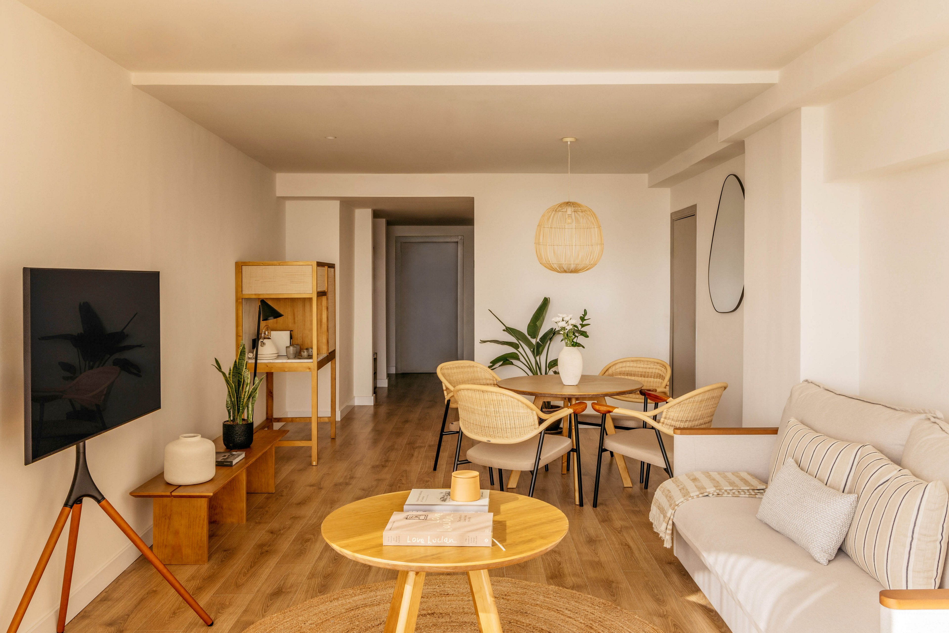 A bright, minimalist living room opens into a small dining area in a modern apartment. Light wood flooring and neutral-toned furniture create a warm, cohesive look throughout the space. A wall-mounted television, round coffee table, and simple shelving unit furnish the living area, while woven chairs surround a wooden dining table. Green plants and soft textiles add natural accents and texture to the clean, contemporary design.