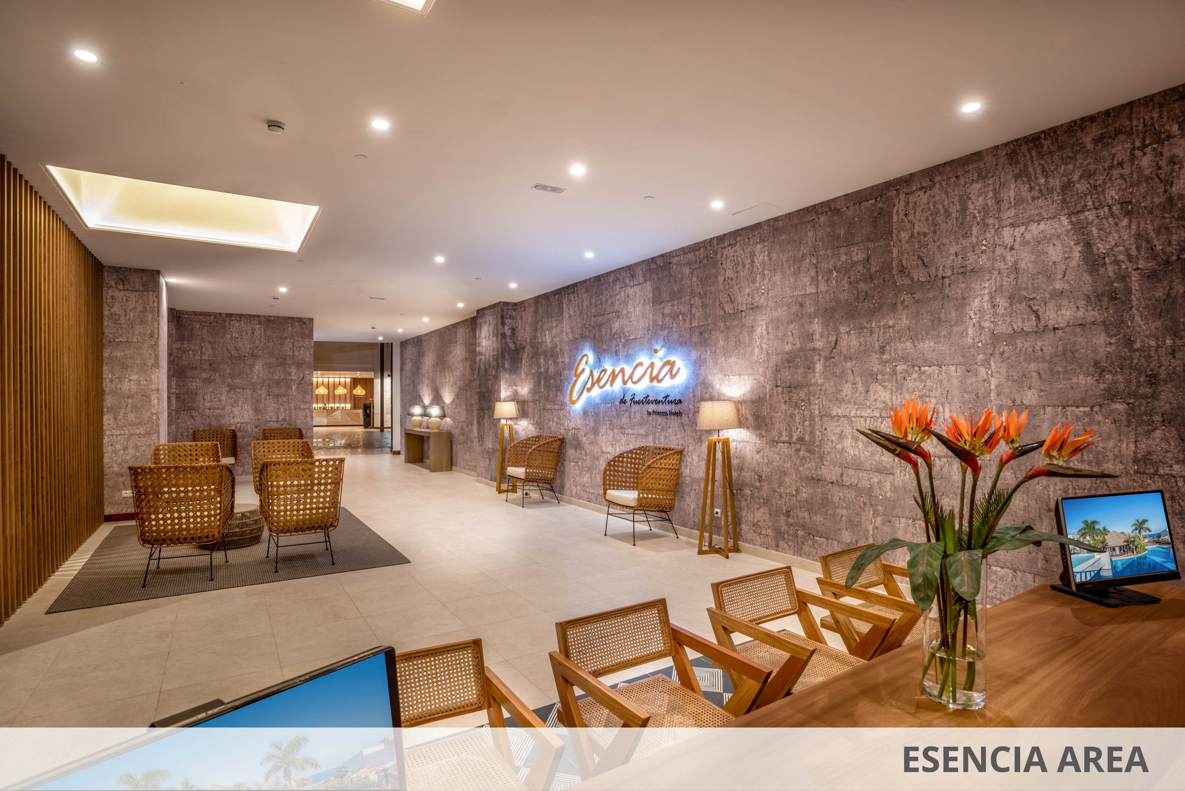 Contemporary hotel lobby space featuring wicker chairs and small tables arranged in cozy seating clusters. Warm recessed lighting and textured walls create an inviting atmosphere. A neon-style wall sign reading "Esencia" adds a focal point in the background. A reception-style desk with tropical flowers and a computer screen is visible in the foreground, with the text "ESENCIA AREA" displayed along the bottom edge of the image.