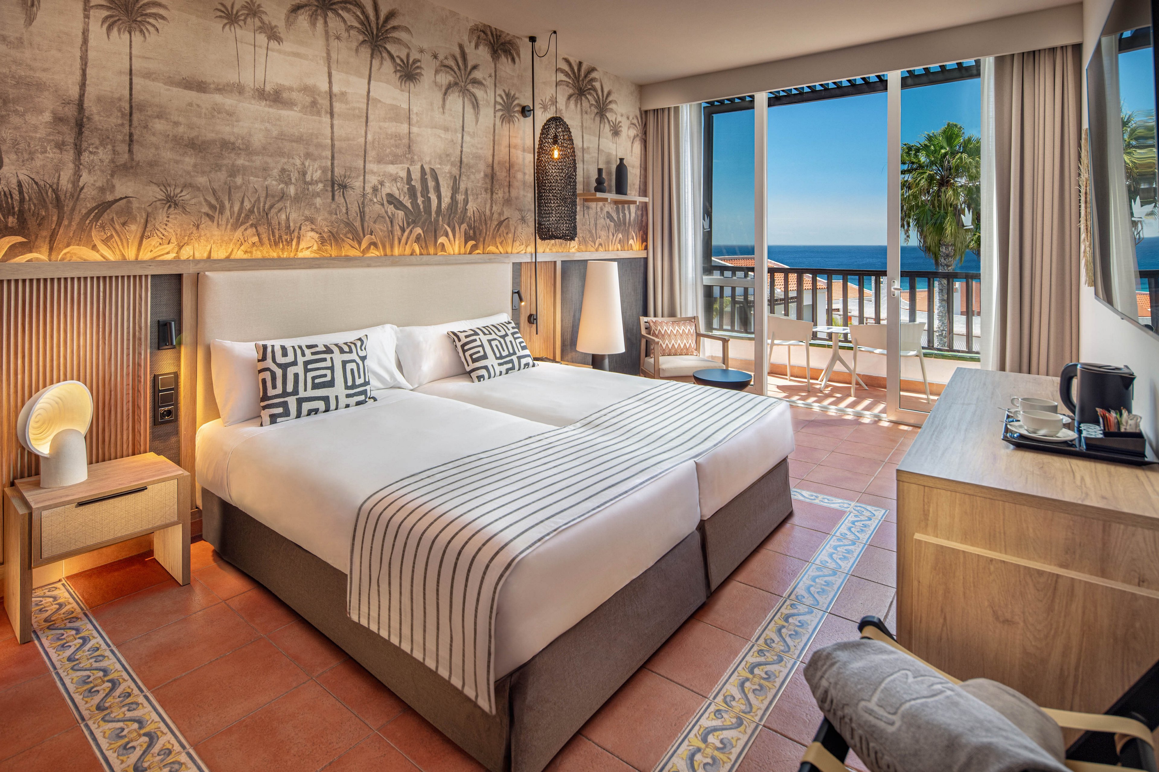 Contemporary hotel room features two neatly made twin beds with striped white bedding and patterned pillows. The space is decorated with a large palm tree mural, warm wood accents, and soft ambient lighting. A desk area with a coffee station, armchair, and floor lamp adds comfort and functionality. Large sliding doors open to a balcony with seating, overlooking the ocean and palm trees under a clear blue sky.