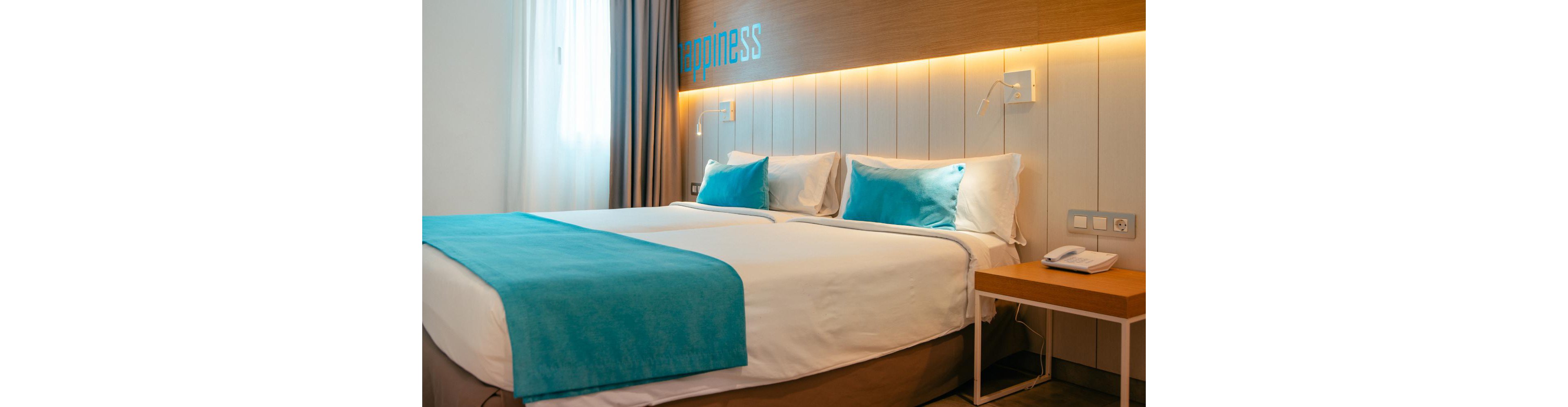 A neatly made double bed with white linens and turquoise pillows and throw blanket stands in a contemporary hotel room. Warm wall lighting illuminates a wooden headboard area with the word "happiness" partially visible above the bed. A bedside table holds a white telephone and sits next to a wall outlet panel. Neutral curtains frame a bright window, adding soft natural light to the clean, minimalist interior. The overall scene conveys comfort, relaxation, and modern hospitality.