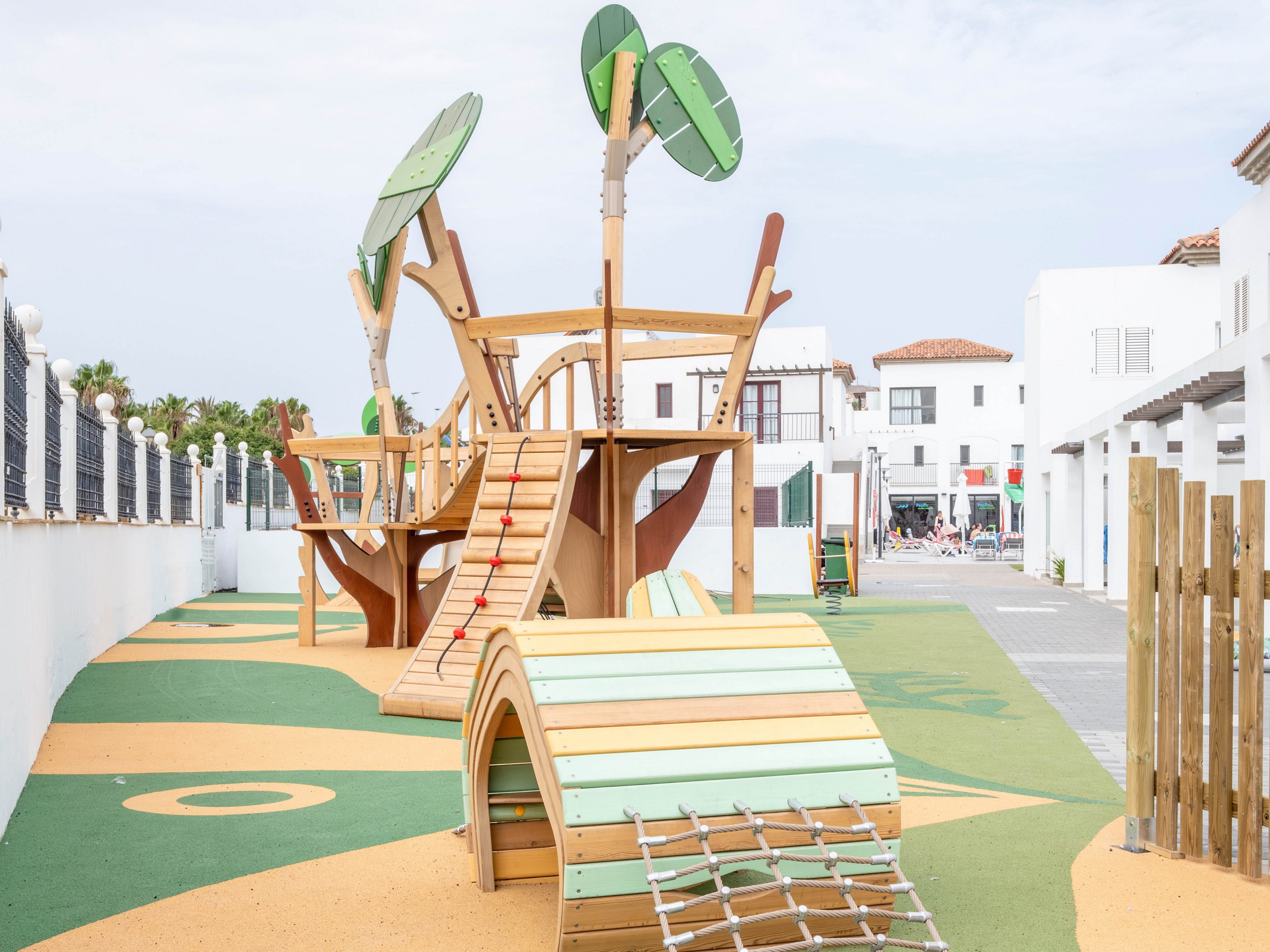 A contemporary wooden playground structure stands in an open outdoor courtyard, featuring slides, climbing nets, and stylized tree elements. The play area is surrounded by white buildings with terracotta roofs, giving a bright Mediterranean feel. Soft, colorful safety flooring with green and yellow shapes covers the ground, and a wooden fence lines one side of the space. The scene is empty of people, emphasizing the clean, well-maintained recreational environment.