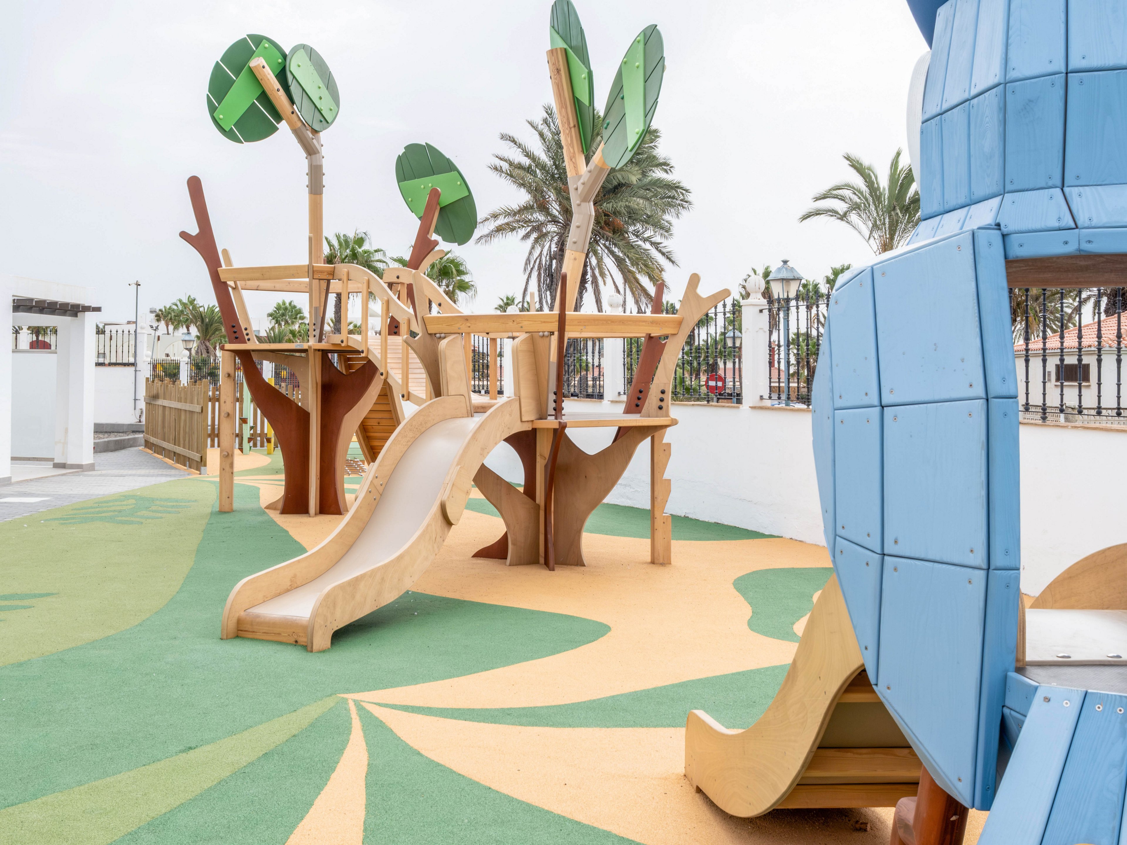 A contemporary outdoor playground features wooden slides and climbing structures with stylized tree elements. The play area is set on a soft, patterned surface in green and yellow tones, suggesting a nature-inspired design. Palm trees and a white wall with railings frame the background, indicating a warm, coastal environment. The scene is bright and inviting, designed for children’s play and recreation.