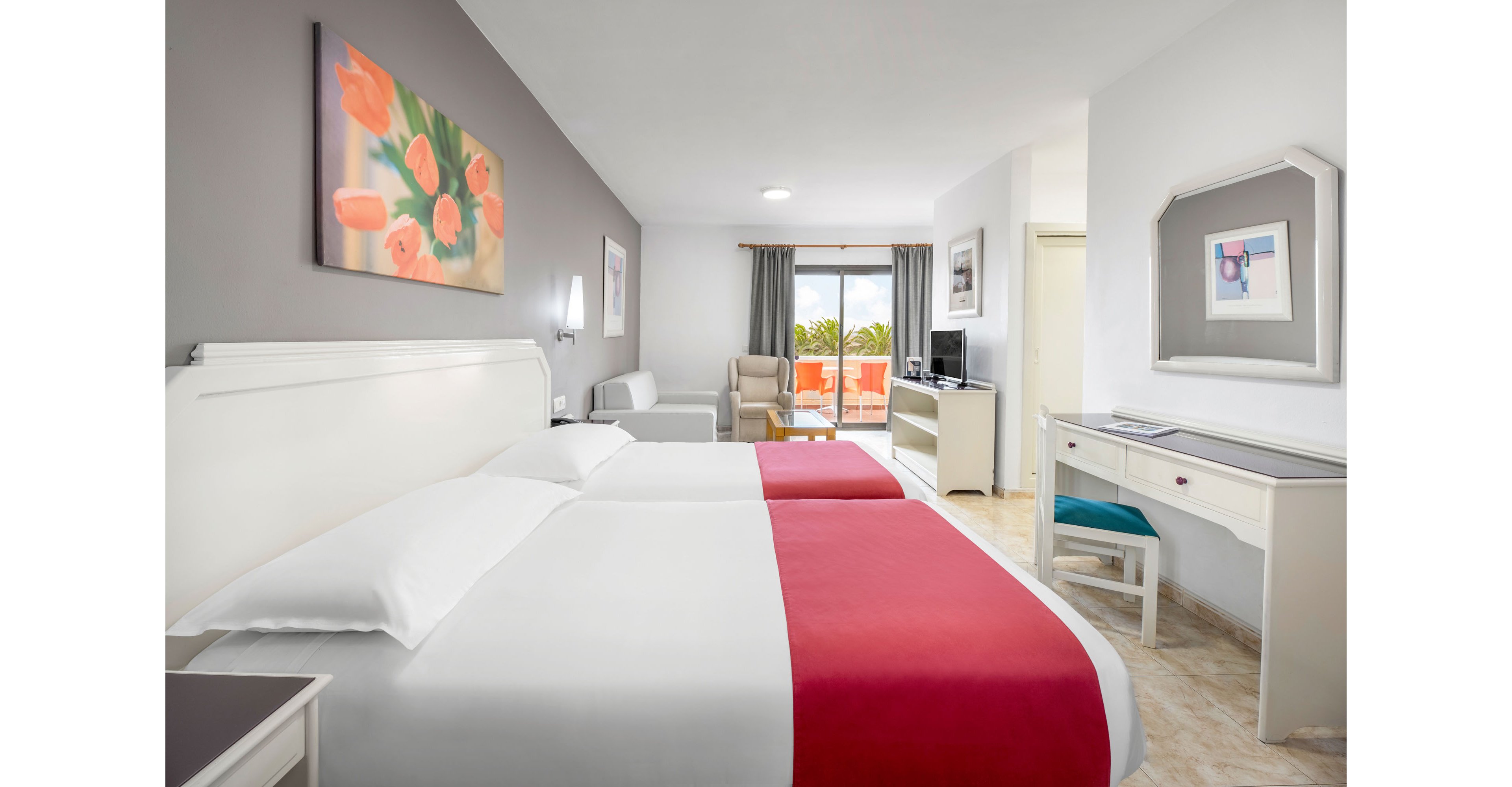 A modern hotel room features two twin beds with white linens and red bed runners against a gray accent wall. The space includes a white desk with chair, a small seating area, and a television on a cabinet. Large glass doors open to a balcony with outdoor chairs and a view of greenery and palm trees. Soft neutral tones, floral artwork, and natural light create a clean, inviting atmosphere.
