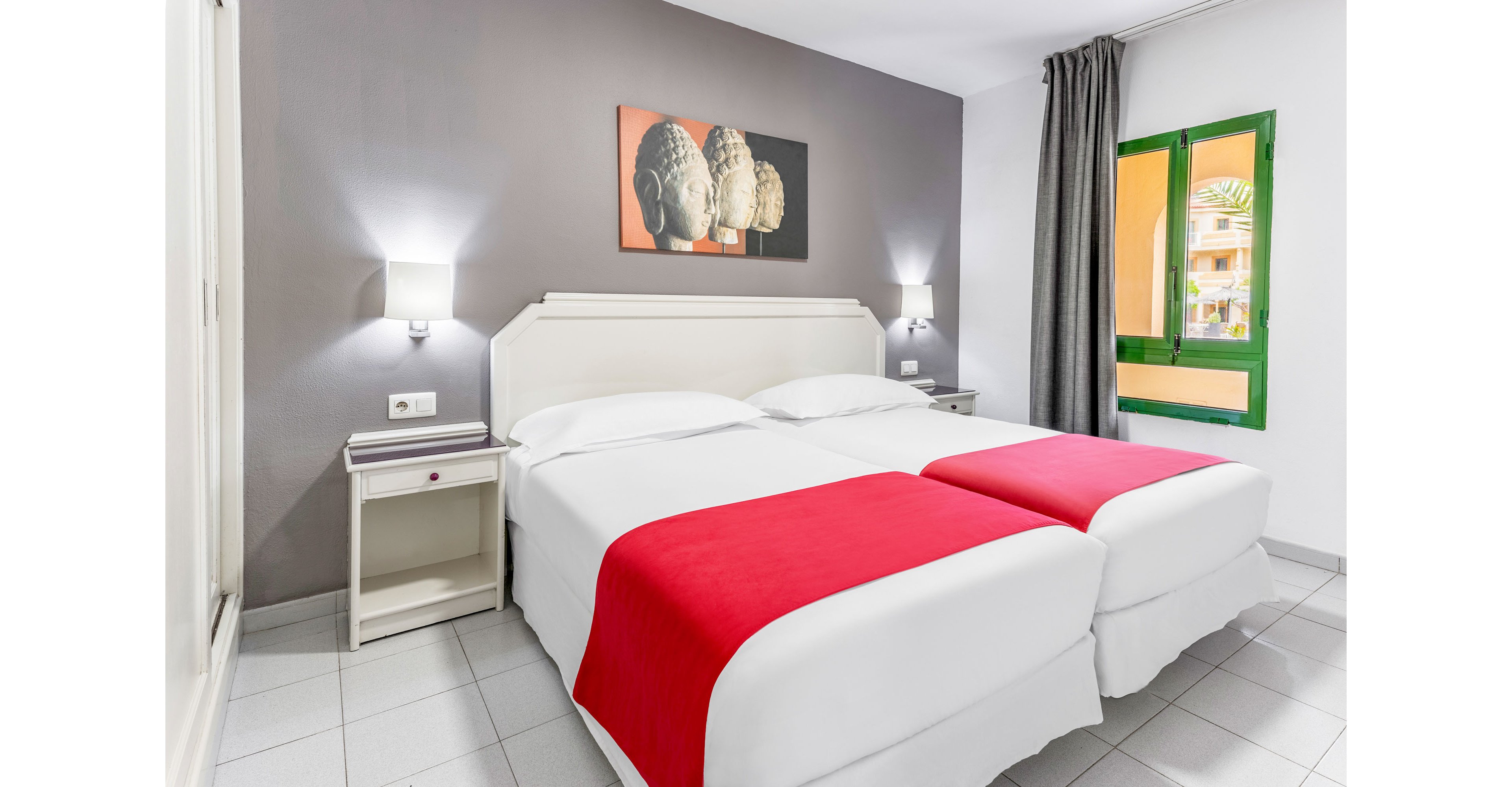 A modern hotel room features two neatly made twin beds with white linens and bold red throws. The space has a gray accent wall, white headboard, and matching bedside tables with wall-mounted lamps. A colorful artwork hangs above the beds, adding visual interest. Natural light enters through a green-framed window with gray curtains, giving the room a bright and welcoming atmosphere.