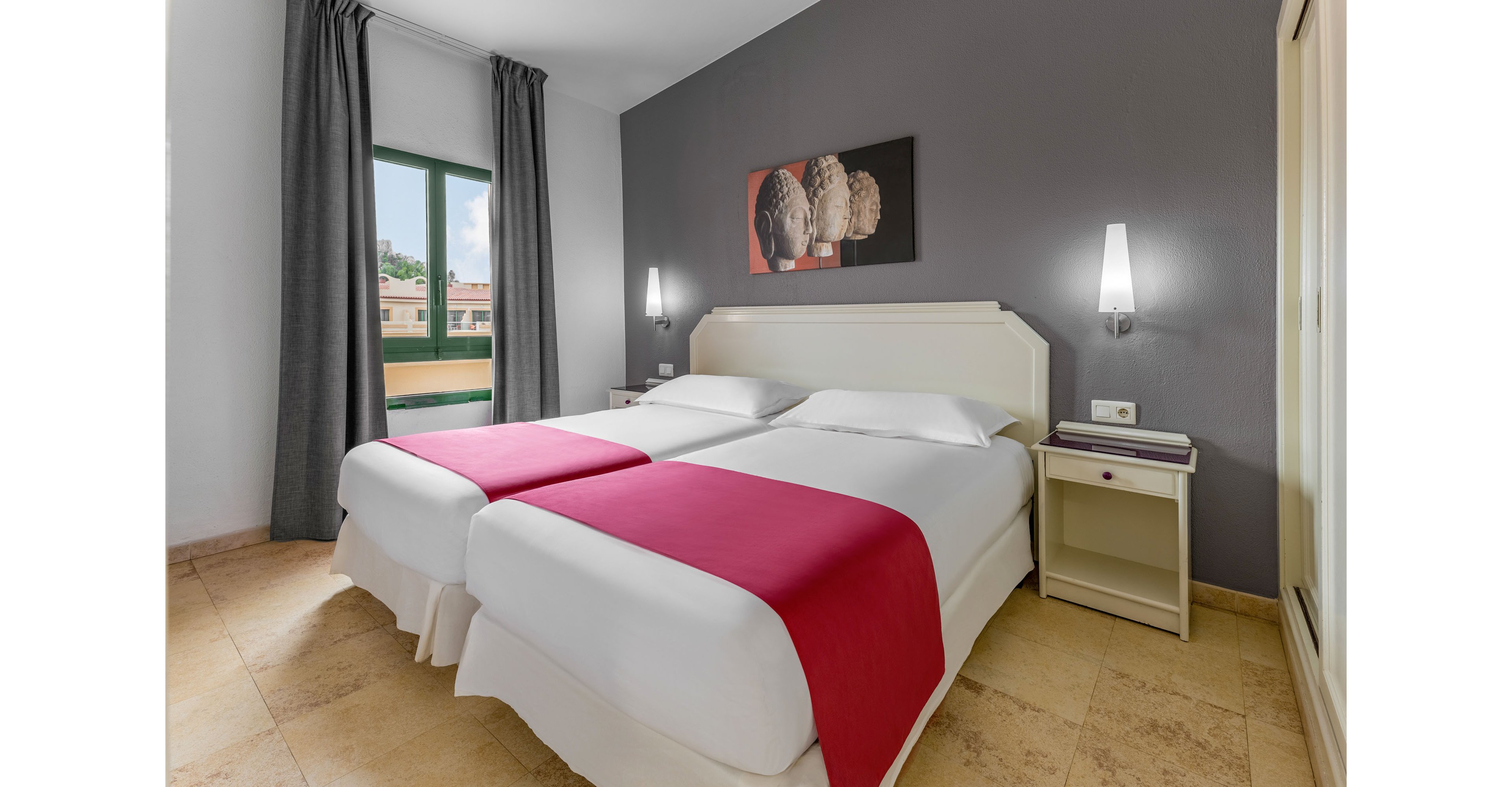 A contemporary hotel room features two neatly made twin beds with white linens and bright red runners. The space includes a gray accent wall, minimalist bedside tables, and wall-mounted lamps providing soft lighting. A large window with dark curtains opens to an outdoor view of neighboring buildings and greenery. A framed artwork above the beds adds a decorative focal point to the clean, modern interior.