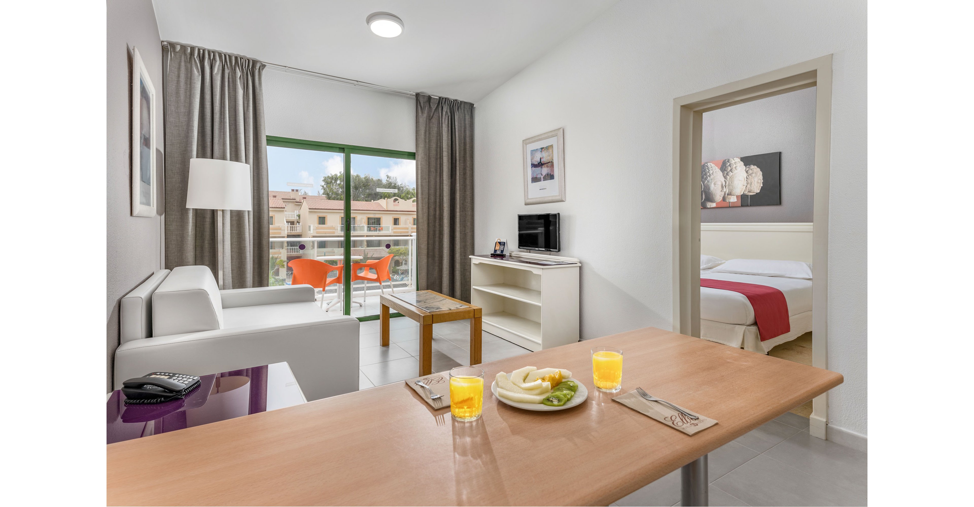 A contemporary hotel suite features a living area with a white sofa, TV, and small dining table. Large sliding glass doors open to a balcony with two orange chairs and a view of neighboring buildings and trees. On the foreground table, two glasses of orange juice and a plate of sliced fruit are neatly arranged beside cutlery. In the background, an adjoining bedroom with a double bed and wall art is visible, creating an inviting vacation atmosphere.