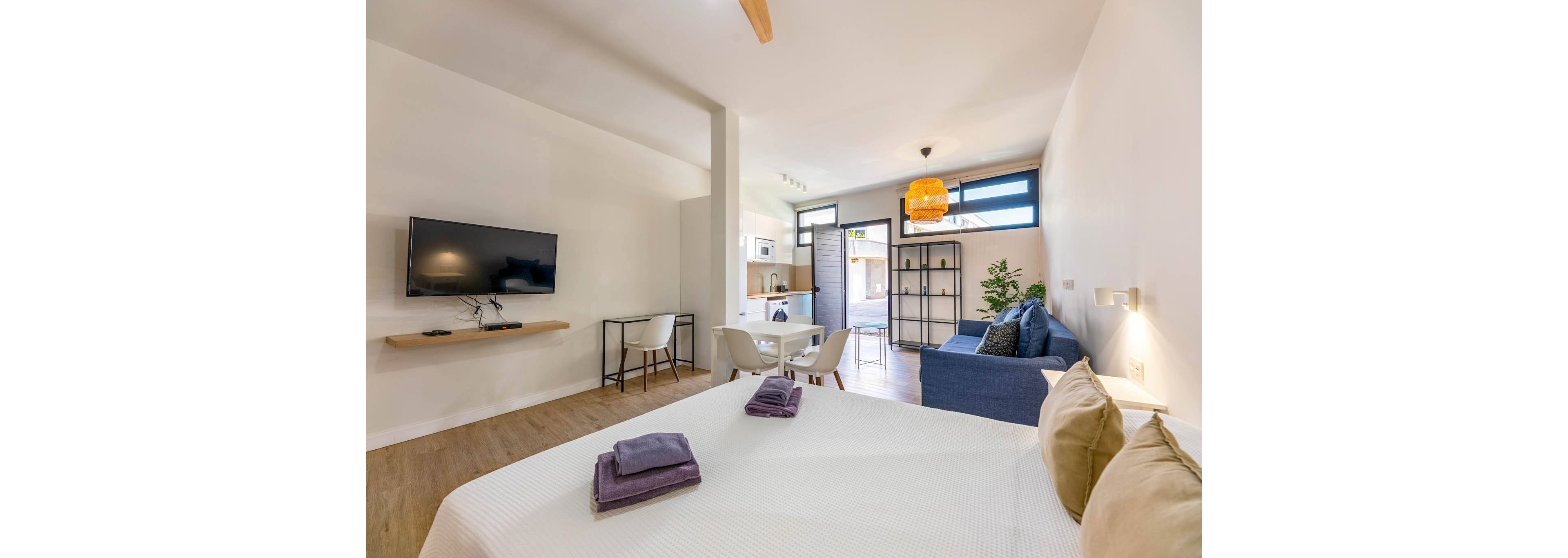 A bright modern studio apartment features a neatly made bed in the foreground with folded towels placed on top. The open-plan space includes a wall-mounted television, a small dining area, and a compact kitchen in the background. A blue sofa and shelving unit sit near large windows that let in natural light, complemented by a warm hanging lamp. Light wood flooring and neutral walls create a clean, minimalist atmosphere suitable for short stays or urban living.