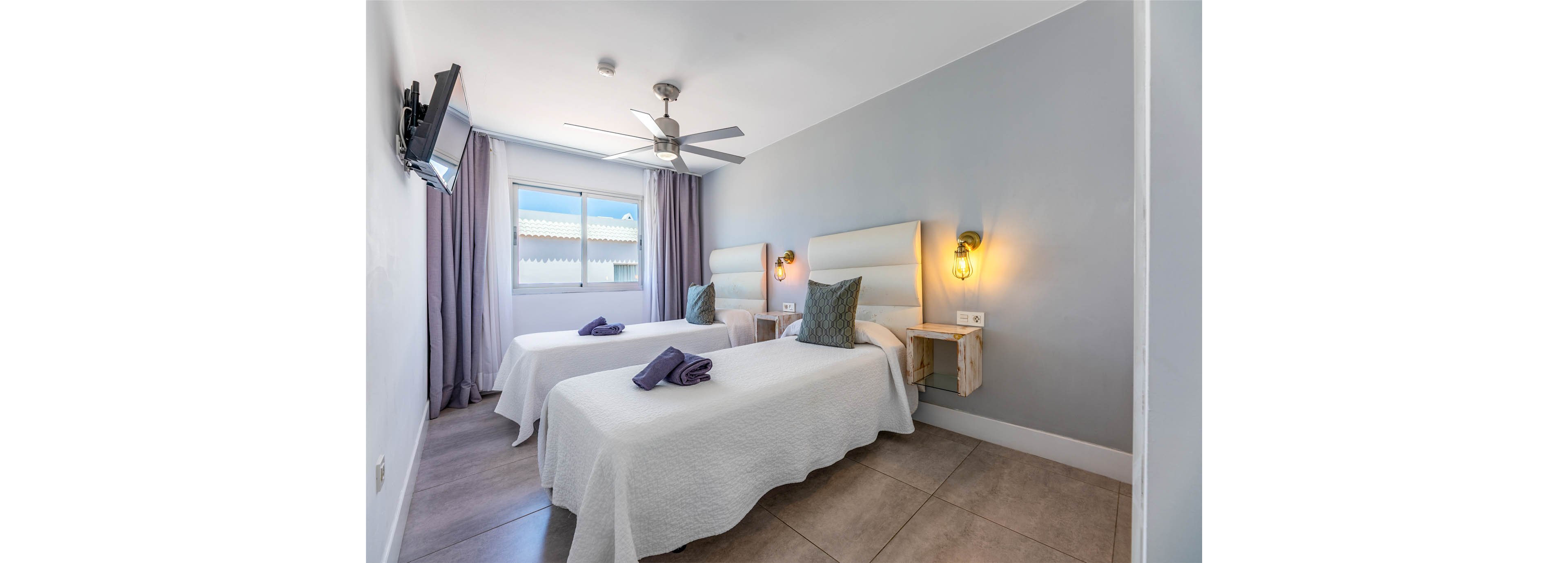A bright modern bedroom features two single beds with white bedding and folded towels placed neatly on top. The room includes a large window with sheer curtains, allowing natural light to illuminate the space. Soft gray walls, minimalist bedside shelves, and warm wall-mounted lights create a clean and inviting atmosphere. A ceiling fan and wall-mounted television add comfort and convenience to the contemporary interior.