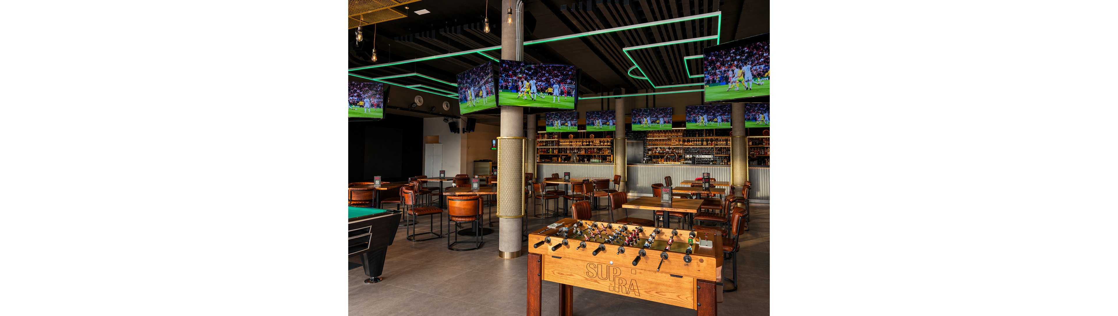 A contemporary sports bar features several large televisions displaying a soccer match, creating an energetic atmosphere. The space includes a foosball table labeled 'SUPRA' and a pool table, surrounded by wooden chairs and tables. Neon ceiling lights add a vibrant touch, while the bar area is stocked with bottles and glasses. The setting is designed for social gatherings and entertainment.