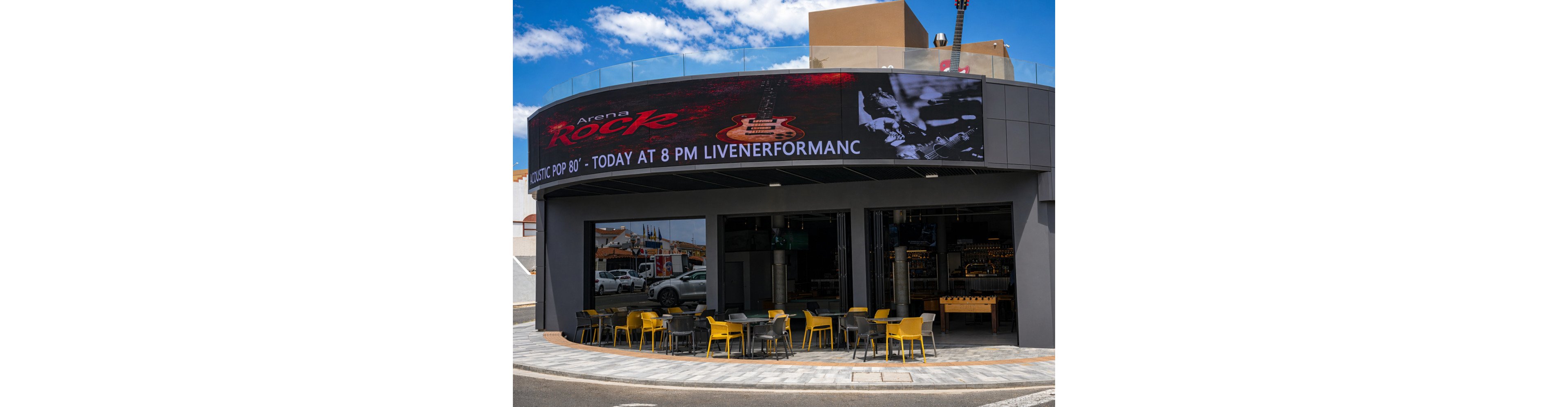 A modern cafe with yellow chairs and tables is situated beneath a large curved digital billboard. The billboard advertises an 'Arena Rock' event, featuring an acoustic pop 80's live performance scheduled for today at 8 PM. The setting is urban, with clear skies and a relaxed outdoor seating arrangement. The visuals include a guitar graphic and partial text on the billboard.
