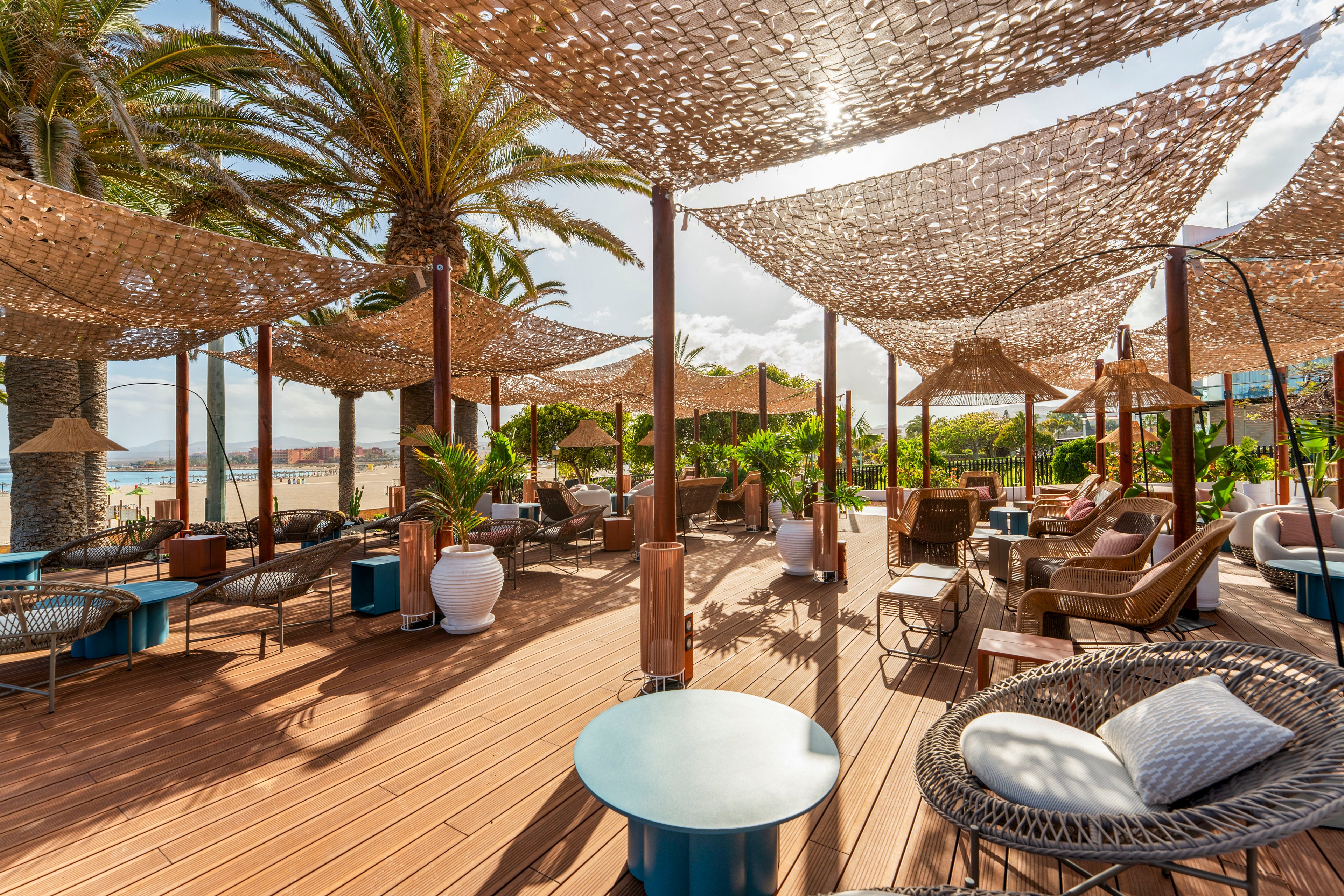 Outdoor lounge area with modern wicker chairs and low tables sits on a wooden deck near a sandy beach. Large palm trees and lush green plants surround the space, creating a tropical atmosphere. Net-style fabric canopies provide dappled shade over the seating area. The scene is bright and sunny, suggesting a relaxed vacation or resort setting.