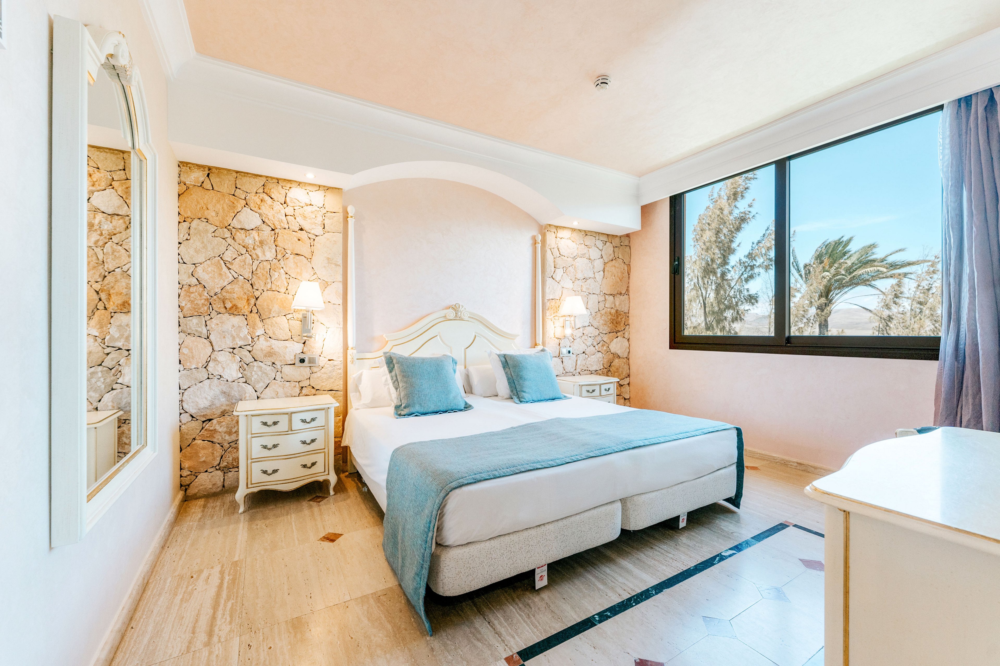 A sunlit hotel bedroom features a neatly made double bed with blue cushions and a light upholstered headboard. Stone accent walls and soft pastel tones create a Mediterranean-inspired interior. Two bedside tables with lamps flank the bed, while a large window reveals palm trees and a clear sky outside. The room feels airy and inviting, with natural light highlighting the clean, minimal decor.