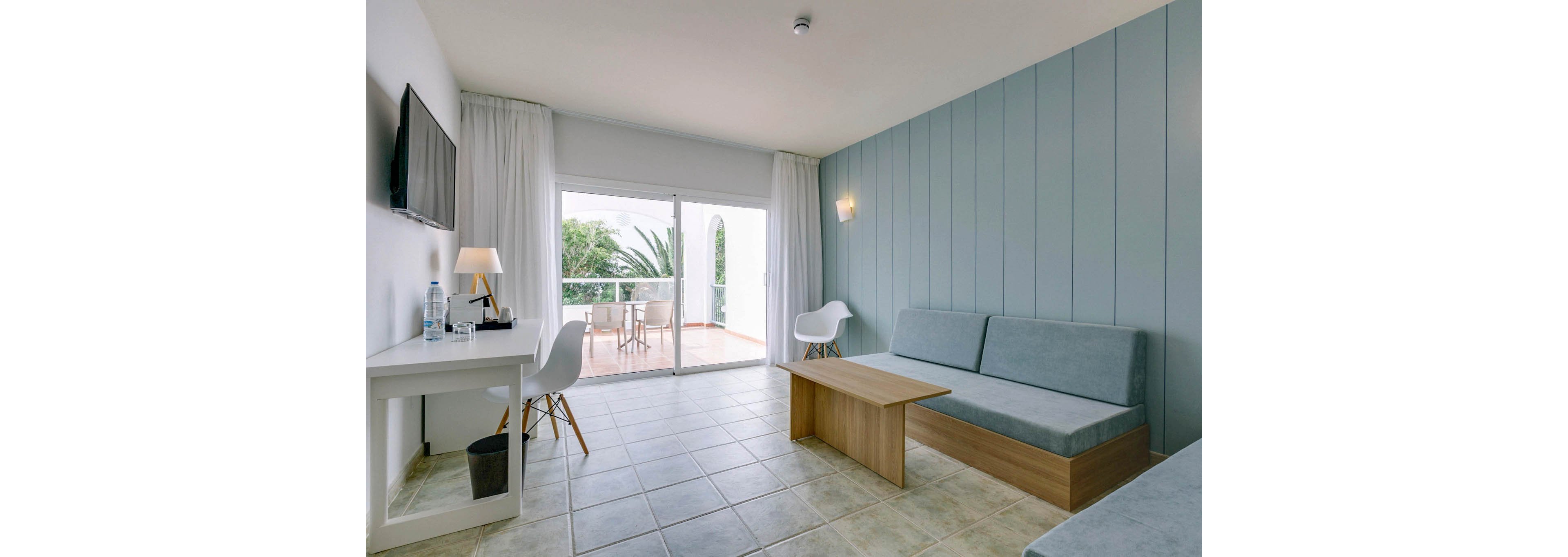 A bright, minimalist hotel room features a light blue accent wall and simple modern furniture. A sofa and wooden coffee table face a wall-mounted television, while a white desk and chair sit near the balcony doors. Large sliding glass doors open to a balcony with outdoor seating and lush greenery visible outside. Soft neutral tones and clean lines create a calm, contemporary atmosphere.
