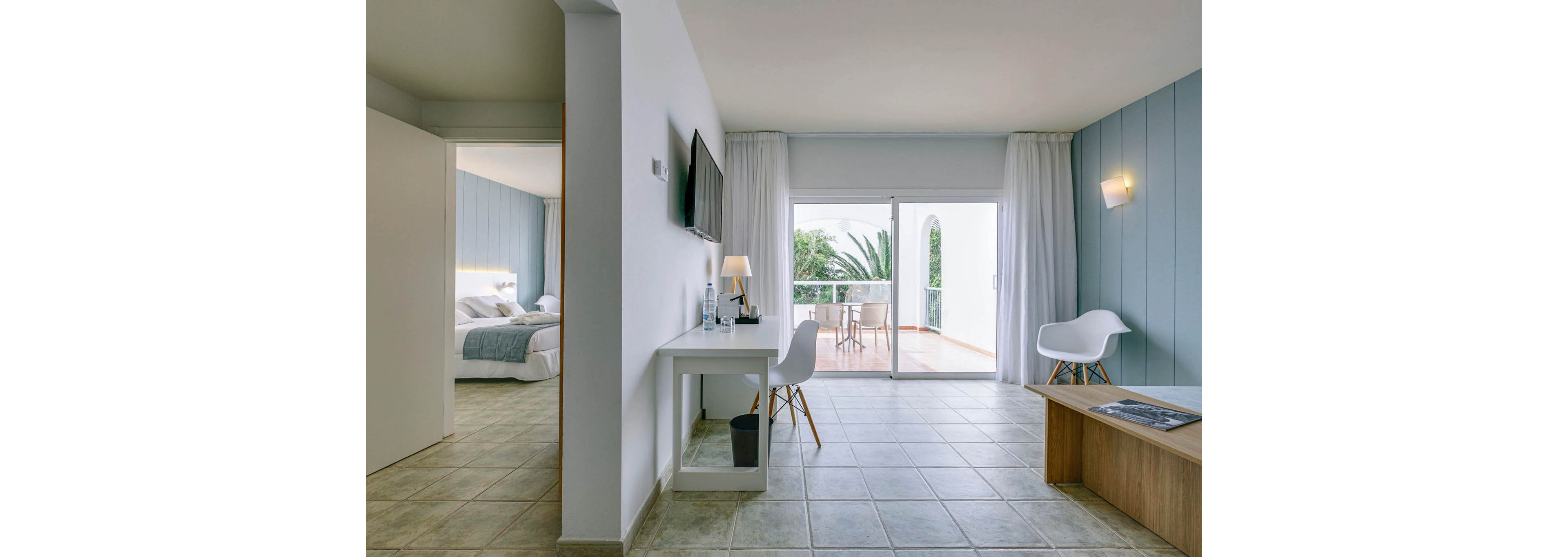 A contemporary hotel suite features a sitting area with a desk, wall-mounted television, and minimalist furniture. Large sliding glass doors open onto a balcony with outdoor chairs and a table, overlooking greenery and palm trees. The adjoining bedroom is visible with neatly made twin beds and soft blue accents. Neutral tiles, white walls, and soft lighting create an airy, relaxing atmosphere.