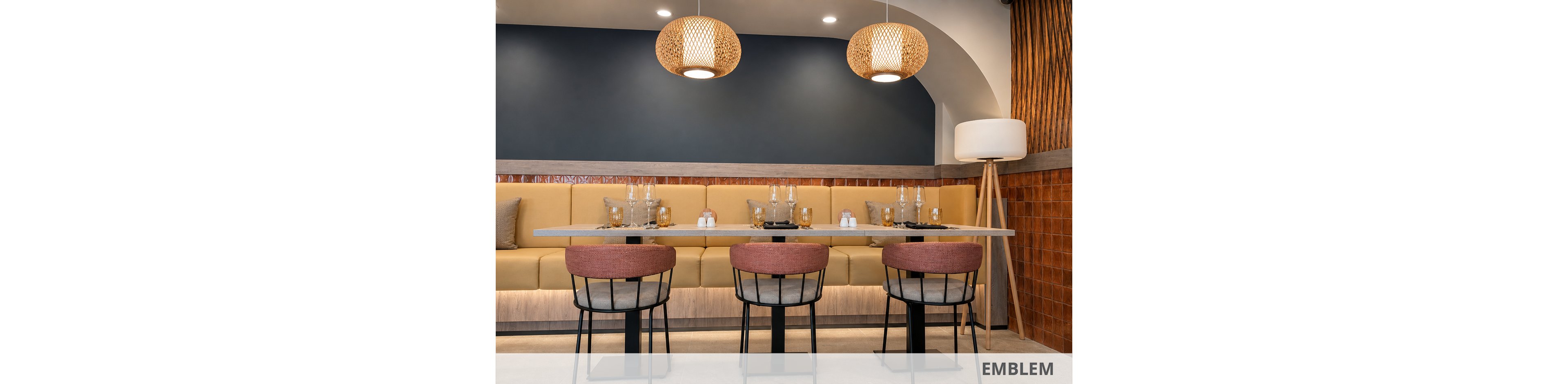 A contemporary restaurant dining area features a long cushioned banquette and four upholstered chairs around sleek tables. The setting is prepared for guests with neatly arranged glassware, plates, and condiments. Warm pendant lights hang from the ceiling, casting a soft glow over the neutral and terracotta color palette. Textured wall tiles and a floor lamp add visual interest and a welcoming, modern ambiance.