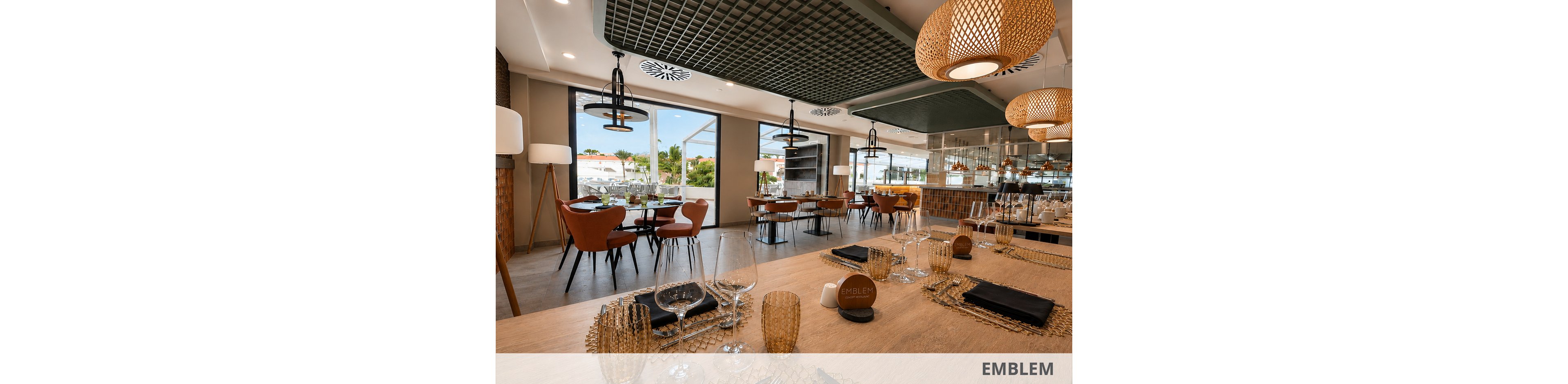 A contemporary restaurant dining room features neatly arranged tables set with glassware, plates, and dark cloth napkins. Warm wooden furniture and woven pendant lights create a cozy yet upscale atmosphere. Large windows reveal an outdoor view with greenery and buildings in the distance. The space appears ready for guests, with no people present and reflections visible in the mirrored wall.