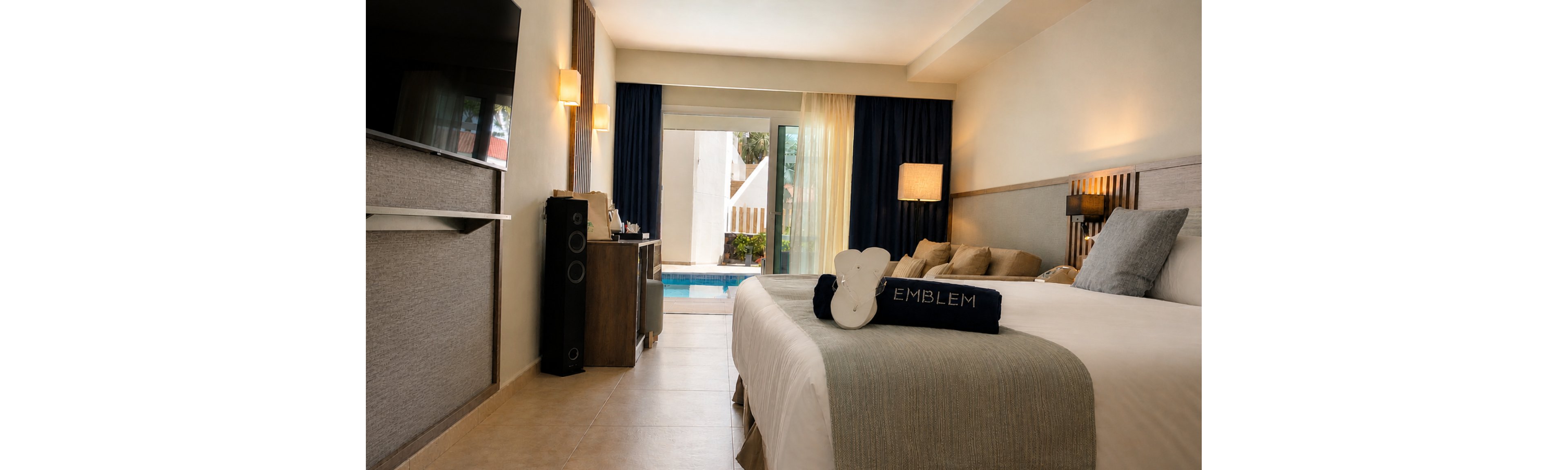 A contemporary hotel room features a neatly made bed with a folded towel labeled "EMBLEM" placed on top. Warm wall sconces and a floor lamp create a cozy ambiance, while a large flat-screen television faces the bed. Sliding glass doors open to a private terrace with direct access to a swimming pool outside. Neutral tones, soft lighting, and minimalist decor emphasize comfort and relaxation.