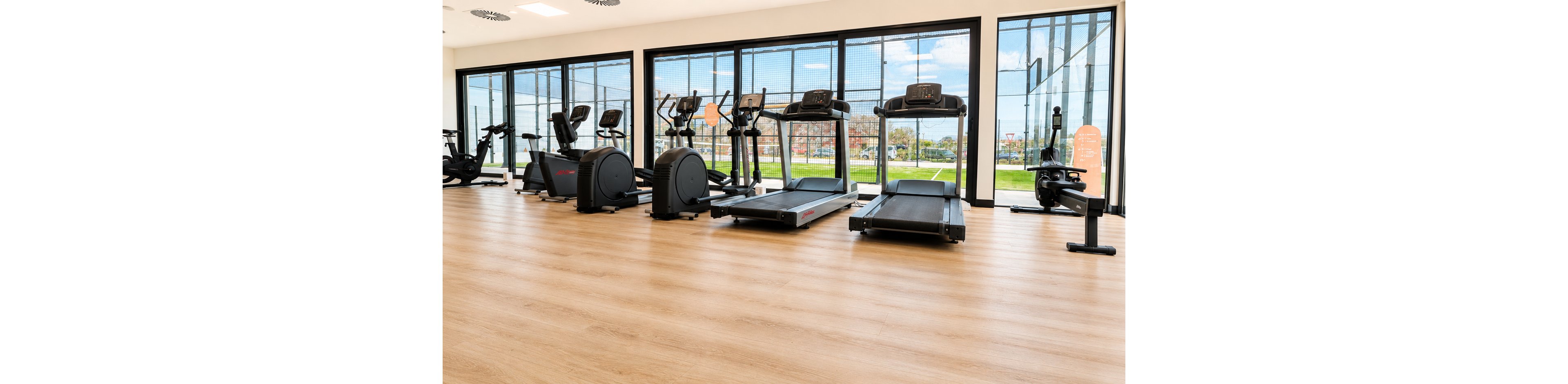 A bright, modern gym features a row of treadmills and elliptical machines lined up on a light wood floor. Large floor-to-ceiling windows let in natural daylight and provide a view of an outdoor sports field and greenery. The space appears clean, organized, and ready for workouts, with no people present. The overall atmosphere is open, minimal, and contemporary.