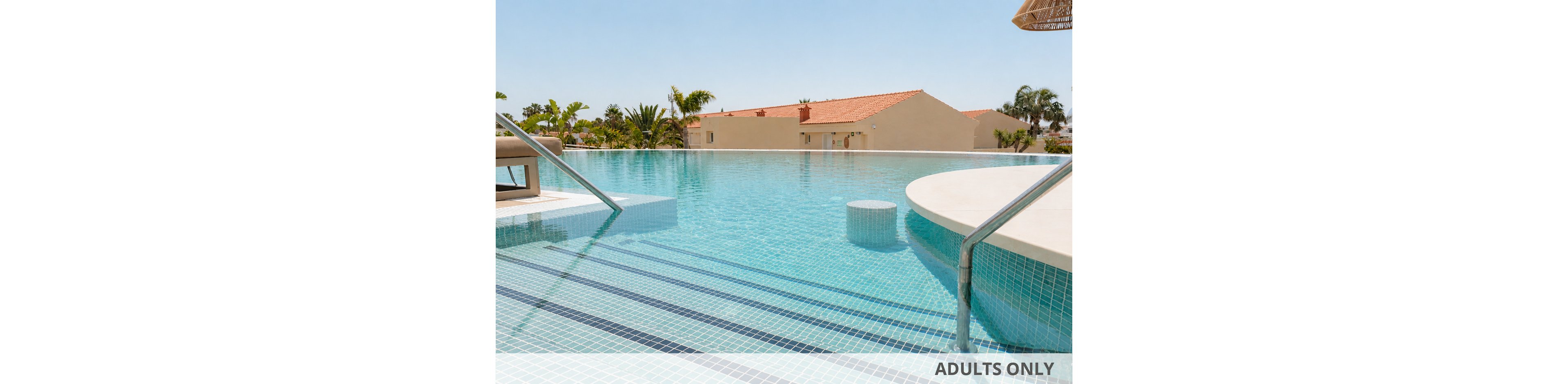 A bright infinity pool stretches toward low resort buildings under a clear blue sky. Stainless steel handrails lead down tiled steps into the shallow water. Palm trees and tropical plants surround the pool area, creating a relaxed vacation atmosphere. A circular in-pool seating area and sun loungers suggest a leisurely, luxurious setting.