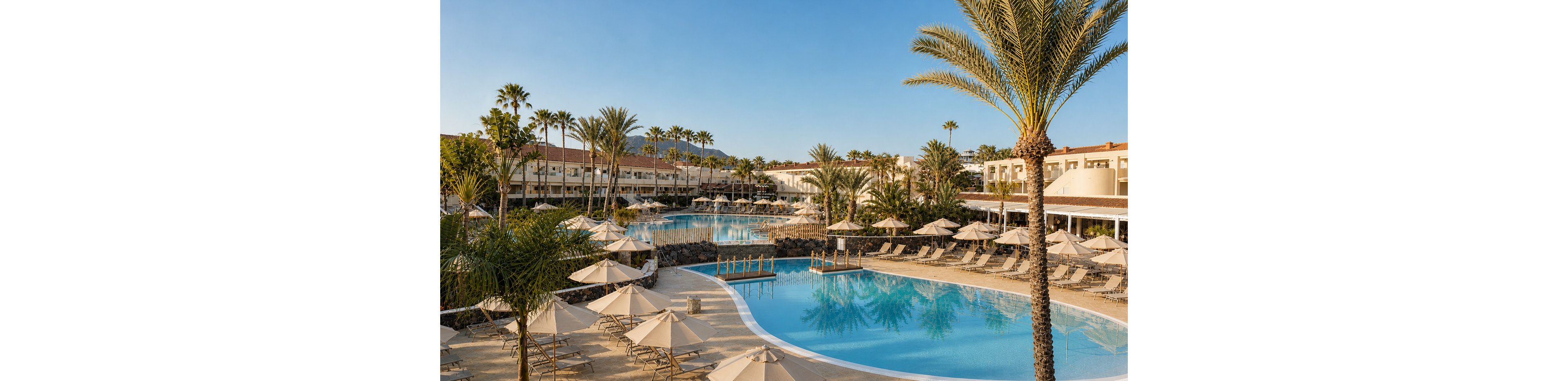 A large resort swimming pool is surrounded by numerous sun loungers and beige umbrellas. Tall palm trees frame the scene, creating a lush tropical atmosphere. Low-rise hotel buildings encircle the pool area, with mountains visible in the distant background. The calm blue water and clear sky suggest a warm, sunny vacation setting.