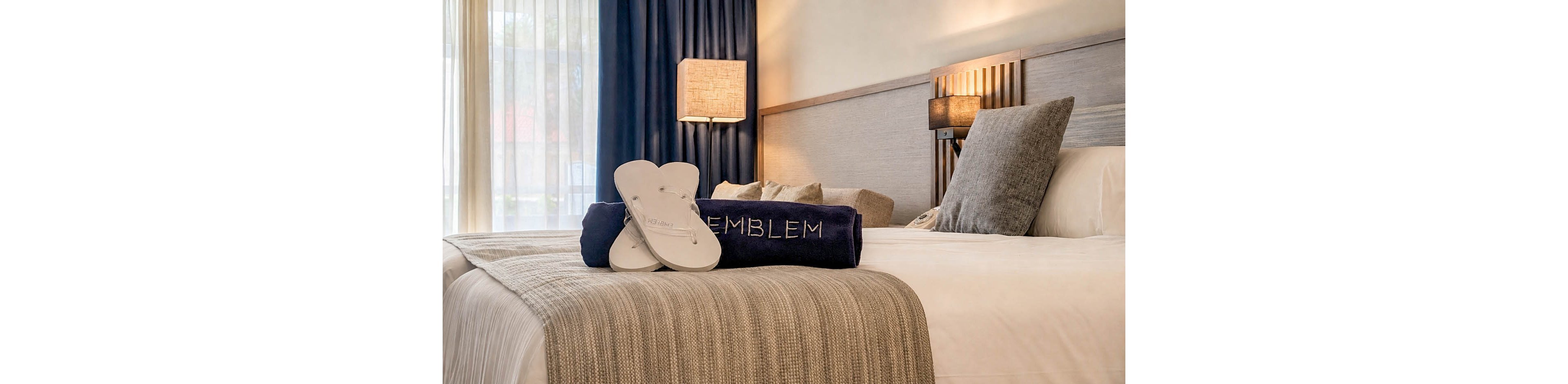 A neatly made hotel bed features a folded blanket and a dark towel with the word EMBLEM visible on it. A pair of white flip-flop slippers rests on top of the towel, suggesting a welcoming guest amenity. Warm bedside lamps and a wooden headboard create a soft, inviting atmosphere. Sheer curtains allow natural light to filter into the modern, comfortable room.