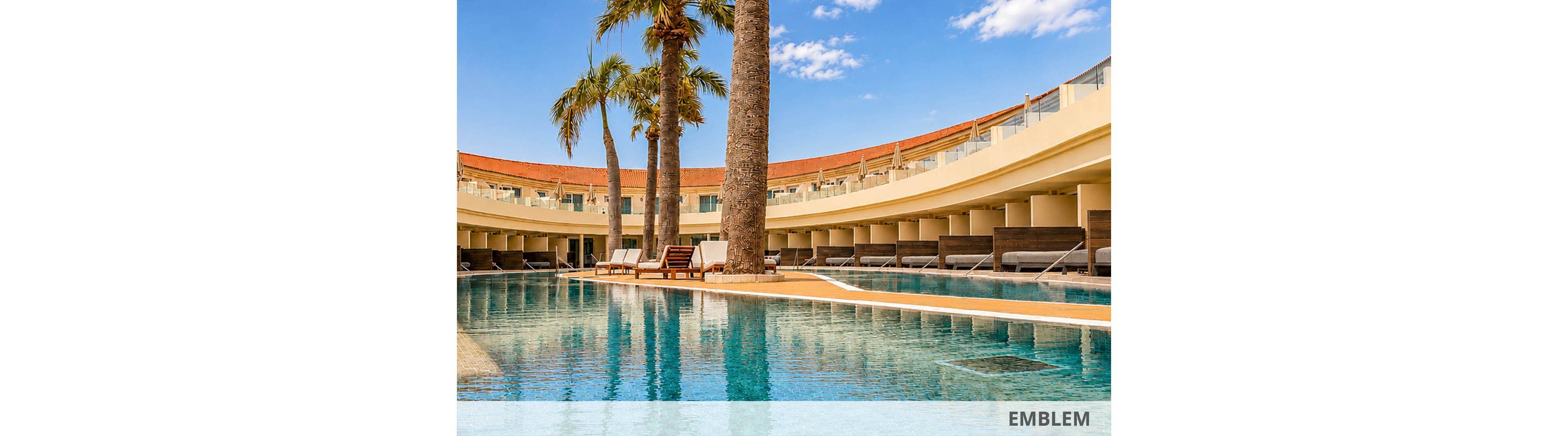 A large, clear swimming pool stretches across the foreground of a modern resort courtyard. Tall palm trees rise from an island in the center of the pool, adding a tropical touch. Curved, multi-level terraces with lounge chairs and shaded daybeds surround the water. The bright blue sky with scattered clouds enhances the sunny, vacation atmosphere.