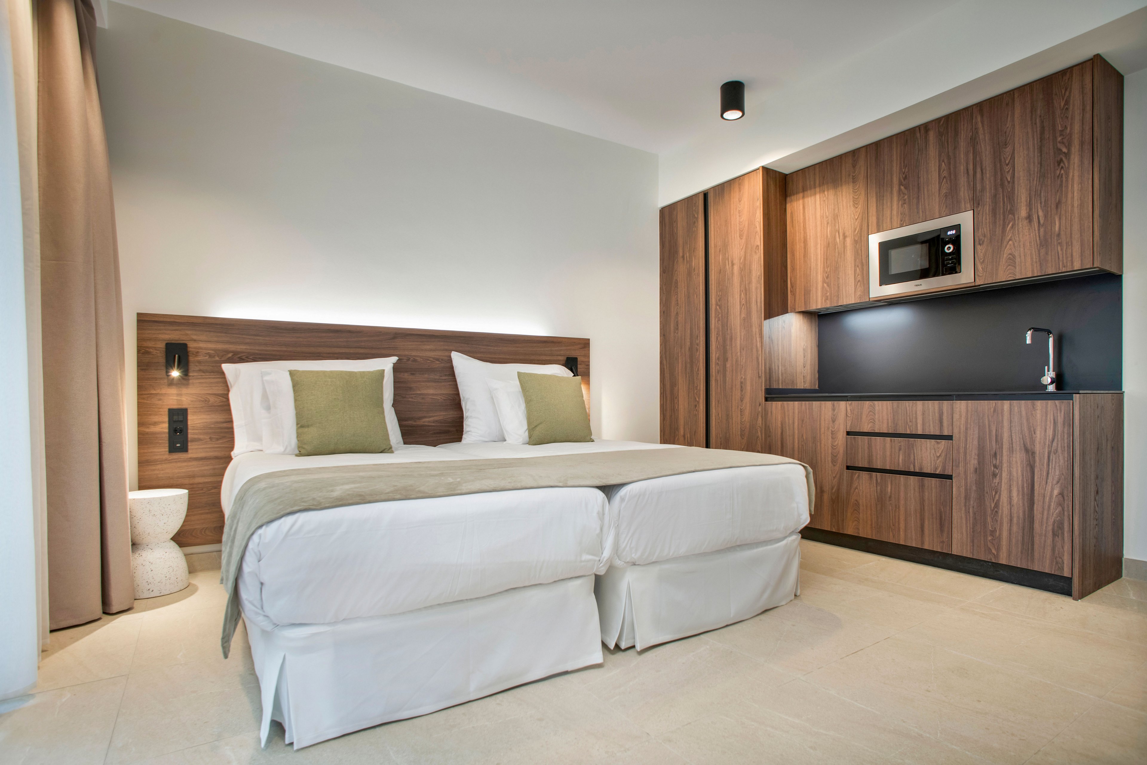 A contemporary hotel room features a double bed with white linens and light green accent pillows. The space includes a built-in wooden headboard and matching cabinetry that integrates a compact kitchenette. A microwave, sink, and storage drawers are neatly arranged along the wall, emphasizing functionality in a small space. Neutral tones, soft lighting, and clean lines create a calm, minimalist atmosphere.