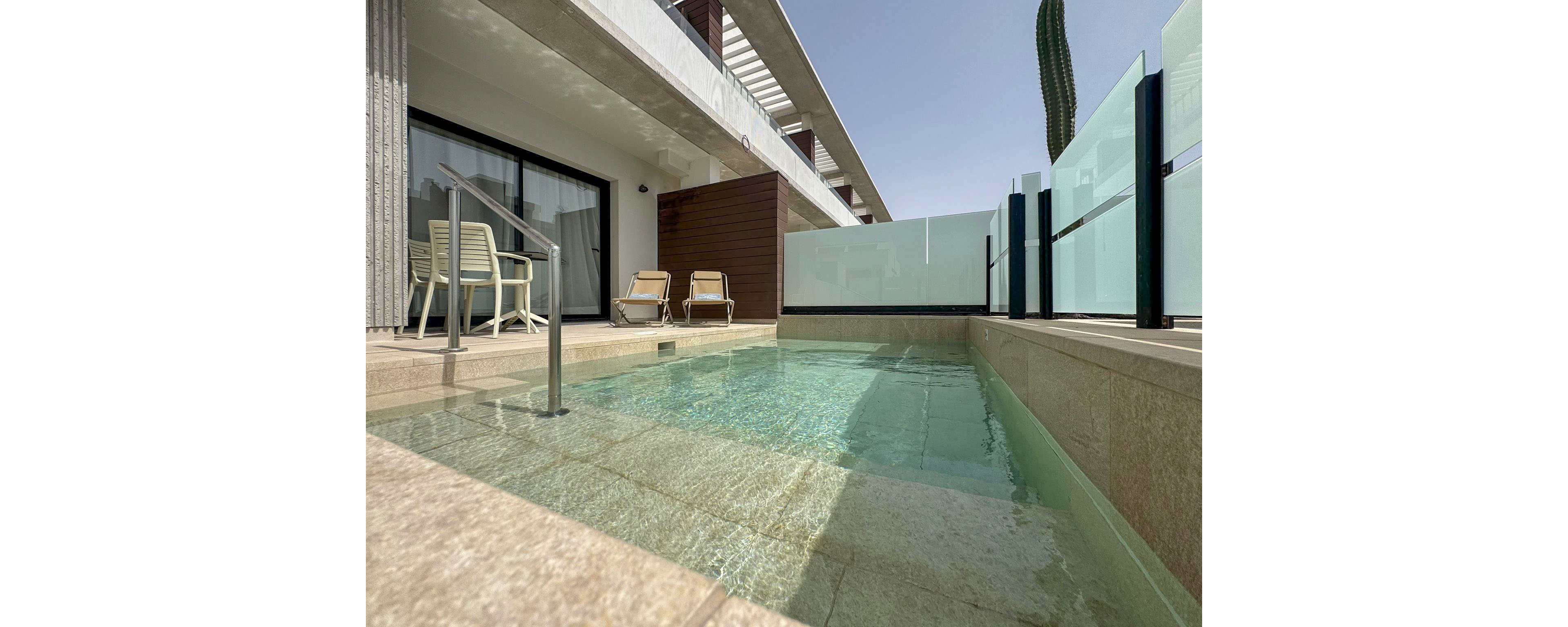 A modern apartment terrace features a shallow private plunge pool with clear water. Two lounge chairs and a small table sit near a sliding glass door leading inside. Frosted glass panels and metal railings provide privacy along the edge of the terrace. The scene is sunlit, with a minimalist architectural style and a tall cactus visible in the background.