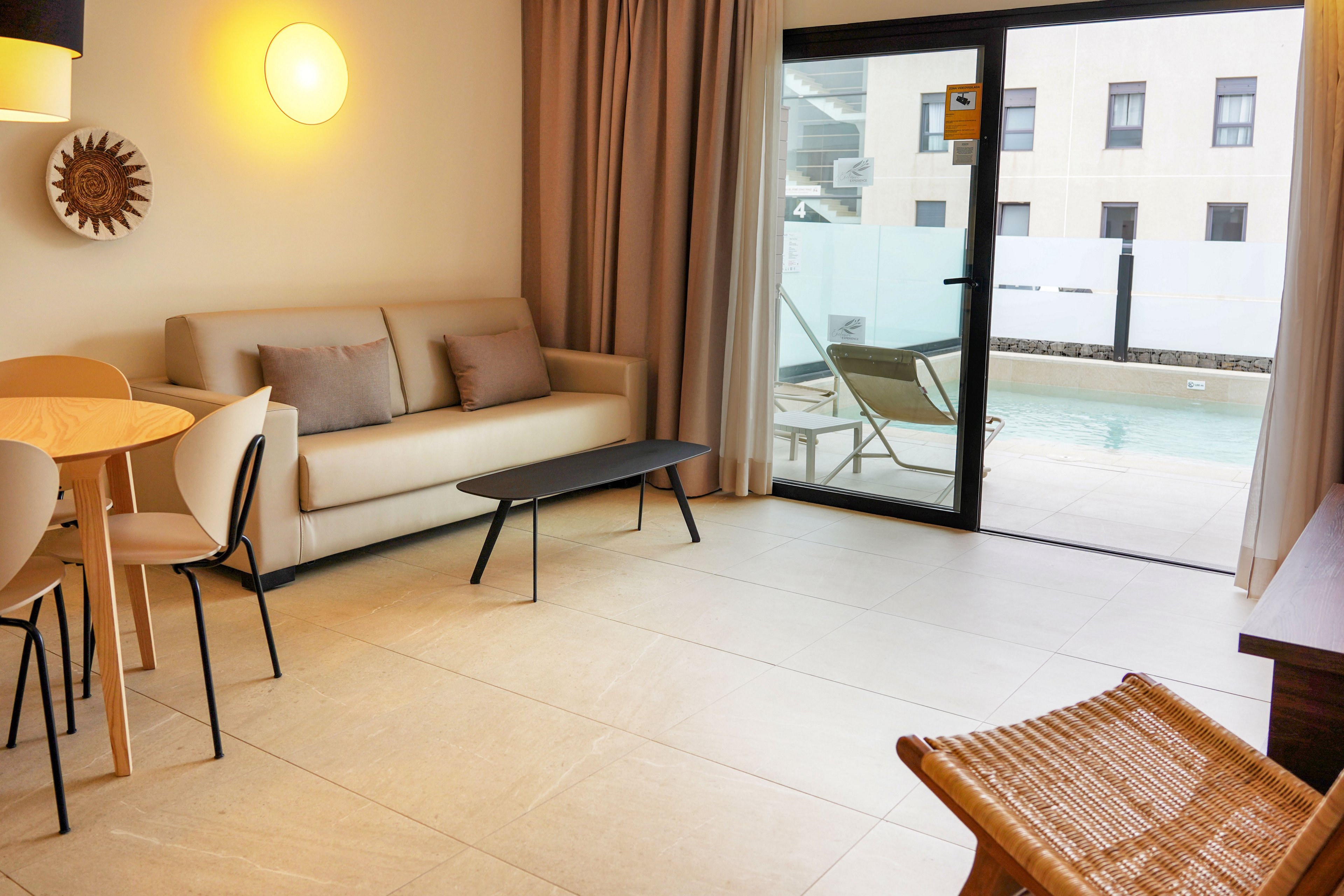 A contemporary living room features a beige sofa with accent pillows and a minimalist black coffee table. To the left, a small round dining table with light wooden chairs sits under a wall-mounted light and decorative sunburst piece. Large sliding glass doors open to a private patio with lounge chairs and a small pool outside. Neutral tones, clean lines, and soft lighting create a calm, modern atmosphere.
