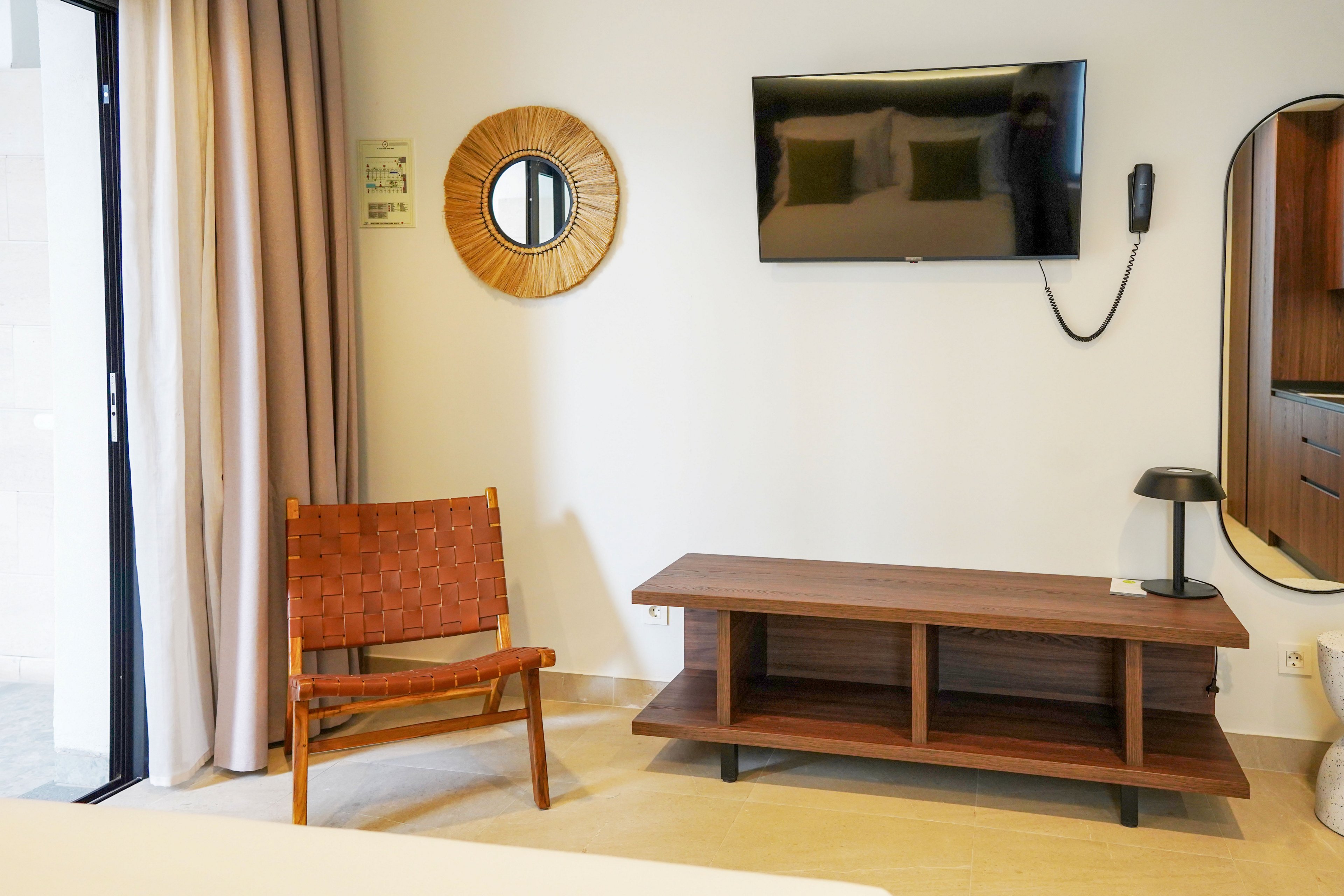 A modern hotel room features a wall-mounted flat-screen television above a low wooden media console. A woven leather chair sits to the left near floor-to-ceiling curtains and a sliding glass door. Decorative elements include a round rattan mirror, a wall-mounted telephone, and a small table lamp on the console. The neutral color palette and clean lines create a calm, minimalist interior atmosphere.