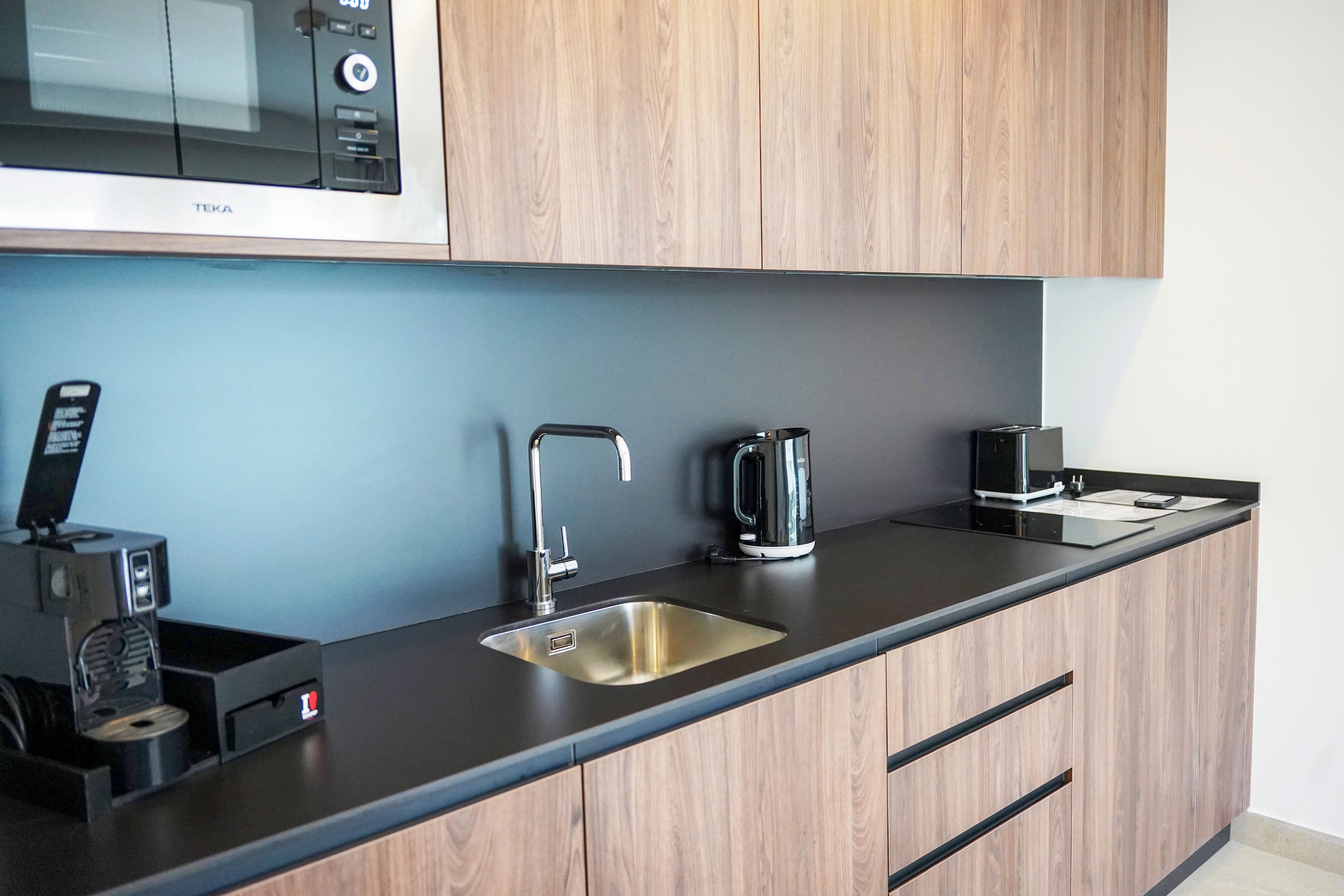 A compact modern kitchenette features wooden cabinets and a black countertop. A stainless steel sink with a sleek faucet is centered against a smooth backsplash. Small appliances, including a coffee machine, electric kettle, toaster, and microwave, are neatly arranged along the counter. The clean, minimal design suggests a contemporary office or apartment kitchen space.