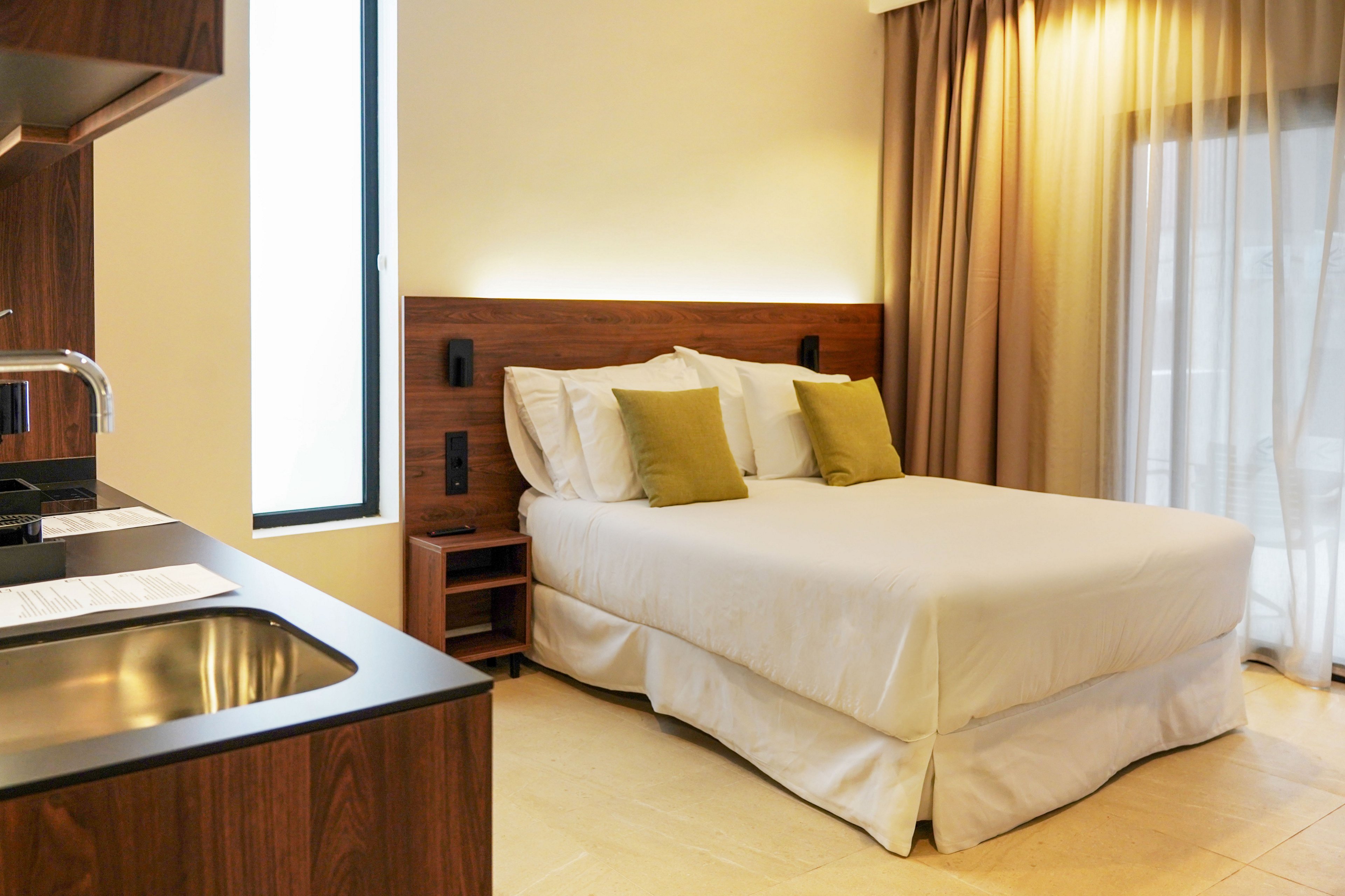 A contemporary hotel-style room features a neatly made double bed with white linens and green accent pillows. The bed is set against a wooden headboard with integrated lighting and wall-mounted fixtures. To the left, a compact kitchenette with a sink and wooden cabinetry adds functionality to the space. Natural light enters through a tall window and a glass door covered with beige curtains, creating a warm and inviting atmosphere.