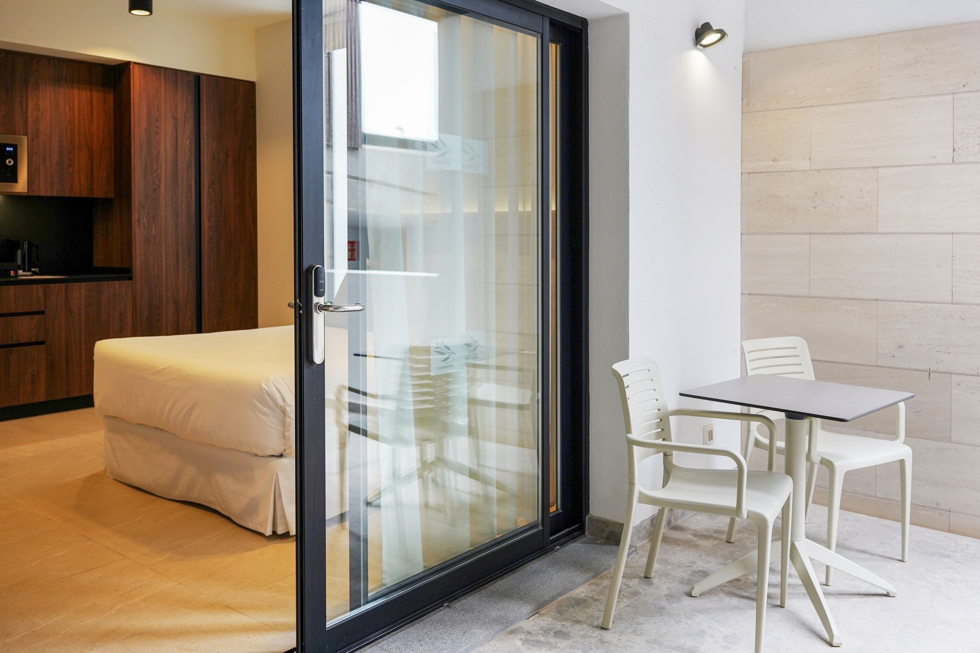 A contemporary hotel room opens onto a small private patio through a large glass sliding door. Inside, a neatly made bed and dark wood cabinetry create a warm, minimalist interior. Outside, a compact table with two chairs sits on a light stone floor against a neutral tiled wall. Soft lighting and clean lines emphasize a calm, modern atmosphere suitable for relaxation or work.