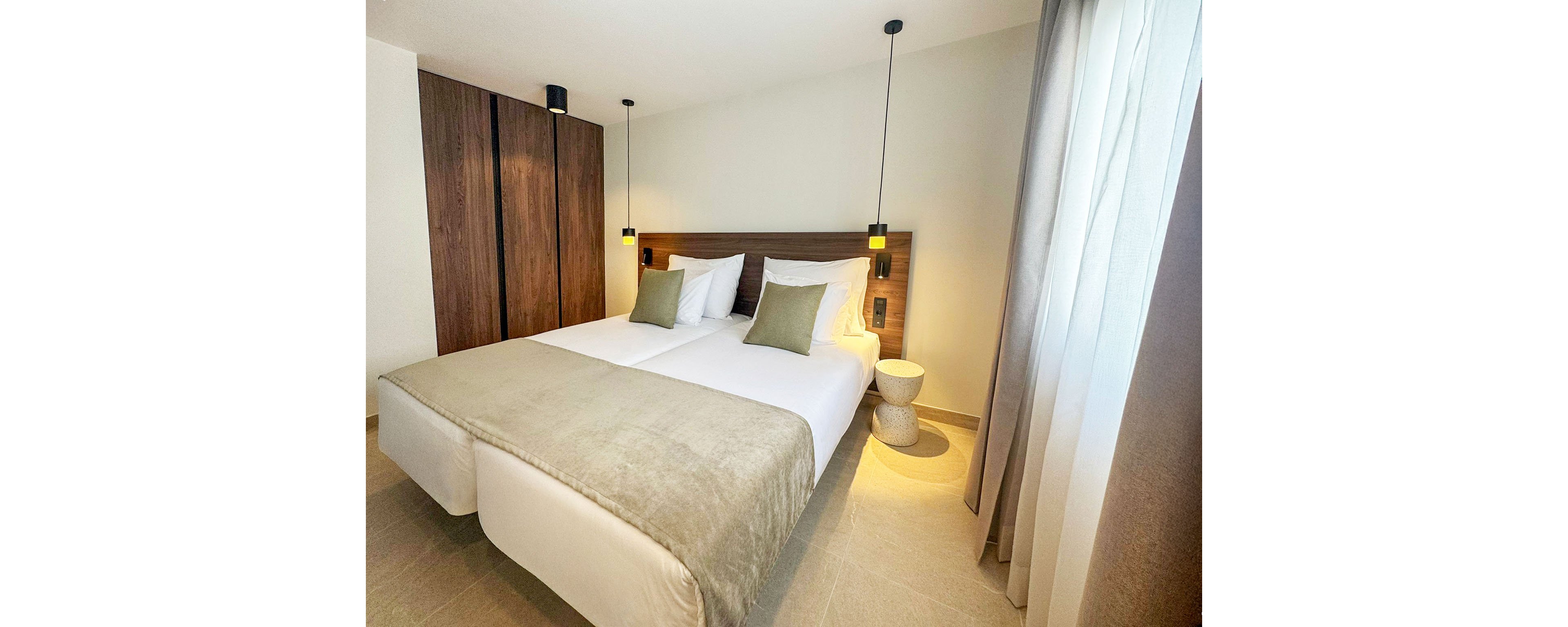 A contemporary hotel bedroom features two neatly made twin beds pushed together with light beige bedding and green accent pillows. The room includes a wooden headboard, matching wardrobe, and minimalist bedside tables. Pendant lights hang above each side of the bed, casting a warm glow. Neutral walls and floor tiles complement the soft natural light coming through sheer curtains, creating a calm and inviting atmosphere.