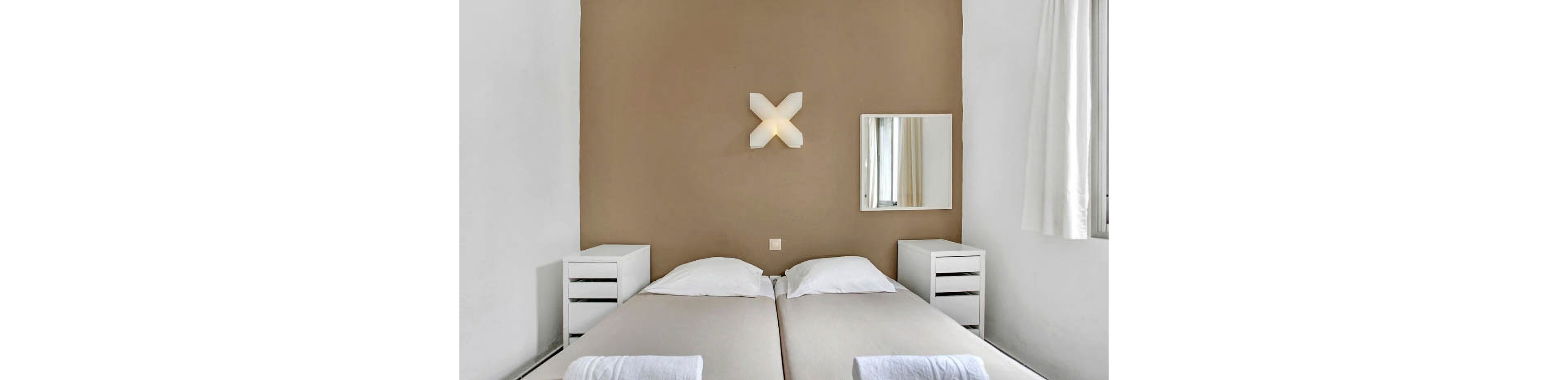 A modern hotel room features two twin beds with white linens, set against a taupe accent wall. The space includes two white nightstands, a wall-mounted mirror, and a unique X-shaped wall light fixture. Natural light enters through a window with sheer curtains, creating a clean and serene atmosphere. The overall palette is neutral, emphasizing simplicity and comfort.