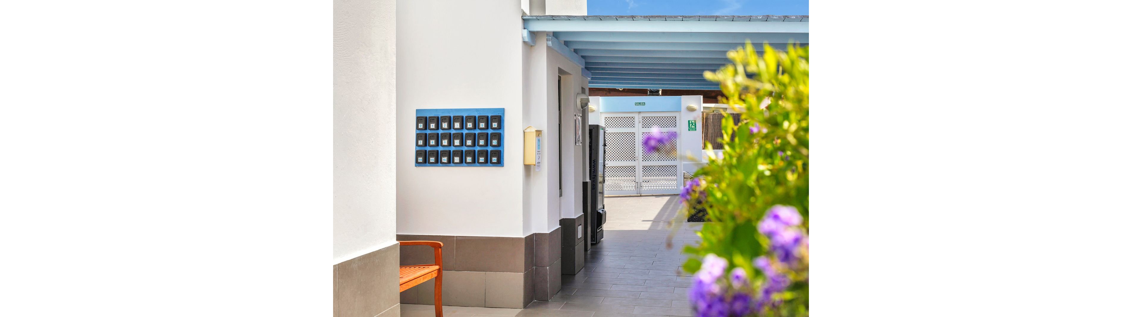 A bright outdoor corridor features a row of blue mailboxes mounted on a white wall. The setting is clean and modern, with a wooden bench and vibrant purple flowers in the foreground. Sunlight casts soft shadows, creating a cheerful and welcoming atmosphere. The corridor leads to a gated area, suggesting a residential or vacation property.