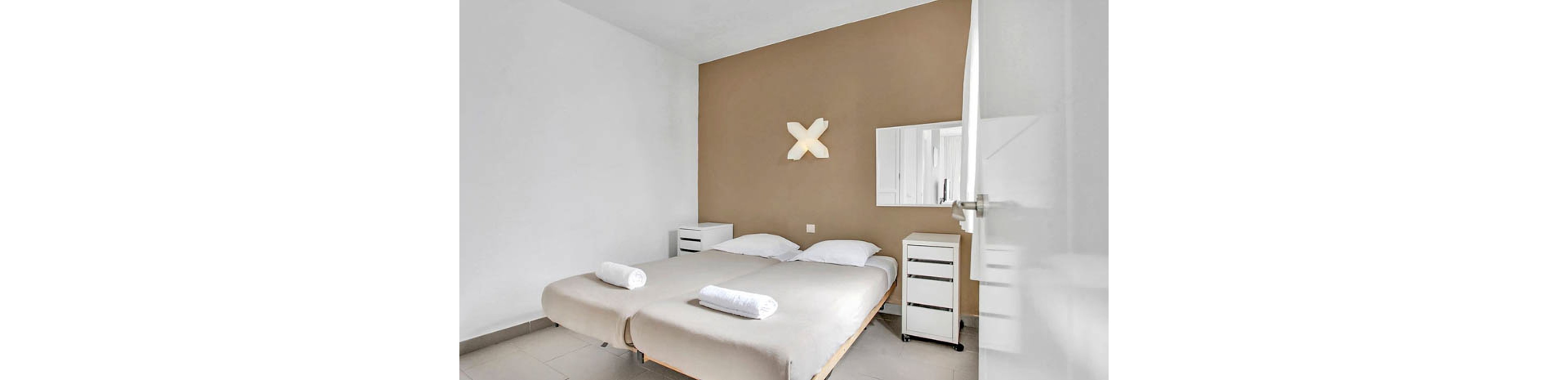 A clean, modern bedroom features two single beds with white linens and rolled towels. The room has a beige accent wall, a geometric wall light, and simple white furniture including nightstands and a small dresser. A wall-mounted mirror adds depth, and the overall style is minimalist with a soft, neutral palette.