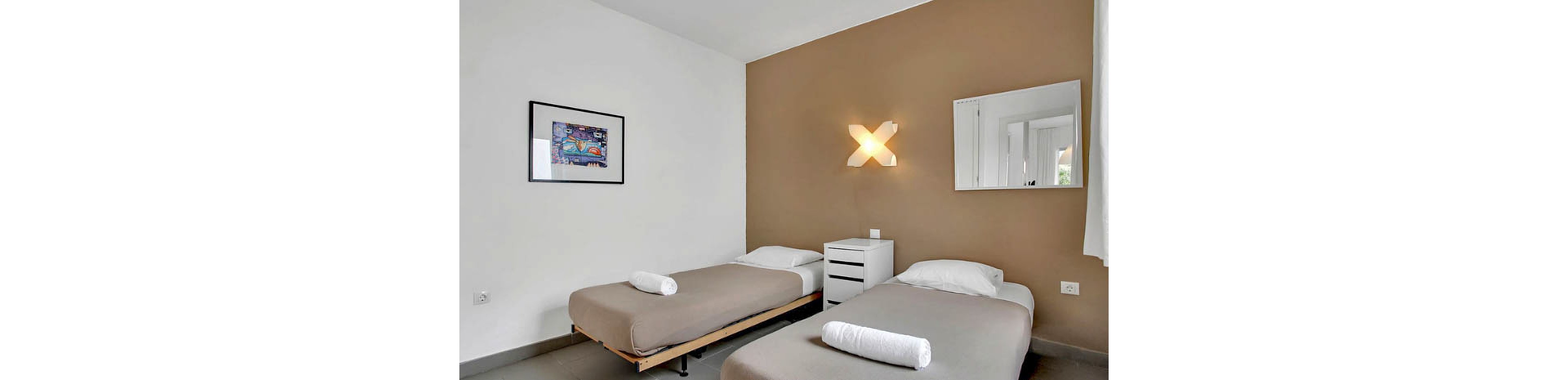 A clean, minimalist bedroom features two single beds with neutral bedding and rolled white towels. The room has a brown accent wall, a unique X-shaped wall light, and a small white nightstand between the beds. A framed artwork and a wall mirror add visual interest, while the overall palette is calm and contemporary.