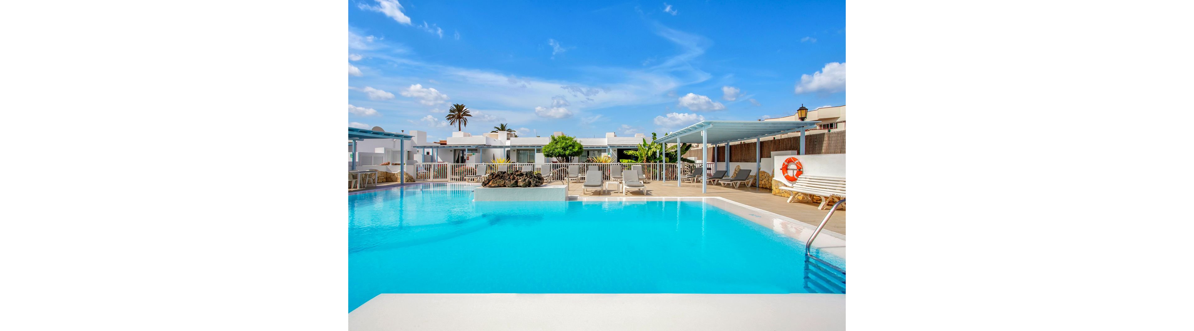 A large, inviting swimming pool is the central focus, surrounded by white modern resort buildings and sun loungers. The setting is outdoors on a sunny day, with a bright blue sky and scattered clouds. A lifebuoy is visible on the poolside fence, and the area is clean and well-maintained. No people are present, emphasizing a tranquil and relaxing atmosphere.