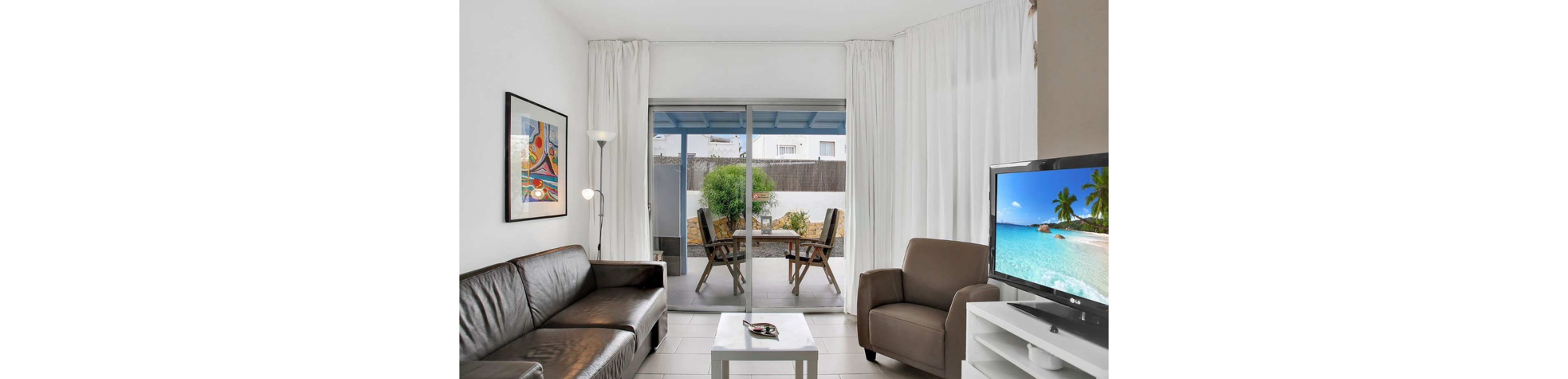 A contemporary living room features a black leather sofa, brown armchair, and a flat-screen television displaying a tropical beach scene. Large glass doors open to a patio with a table and chairs, letting in natural light. The space is decorated with a colorful abstract painting, white curtains, and a minimalist coffee table, creating a clean and inviting atmosphere.